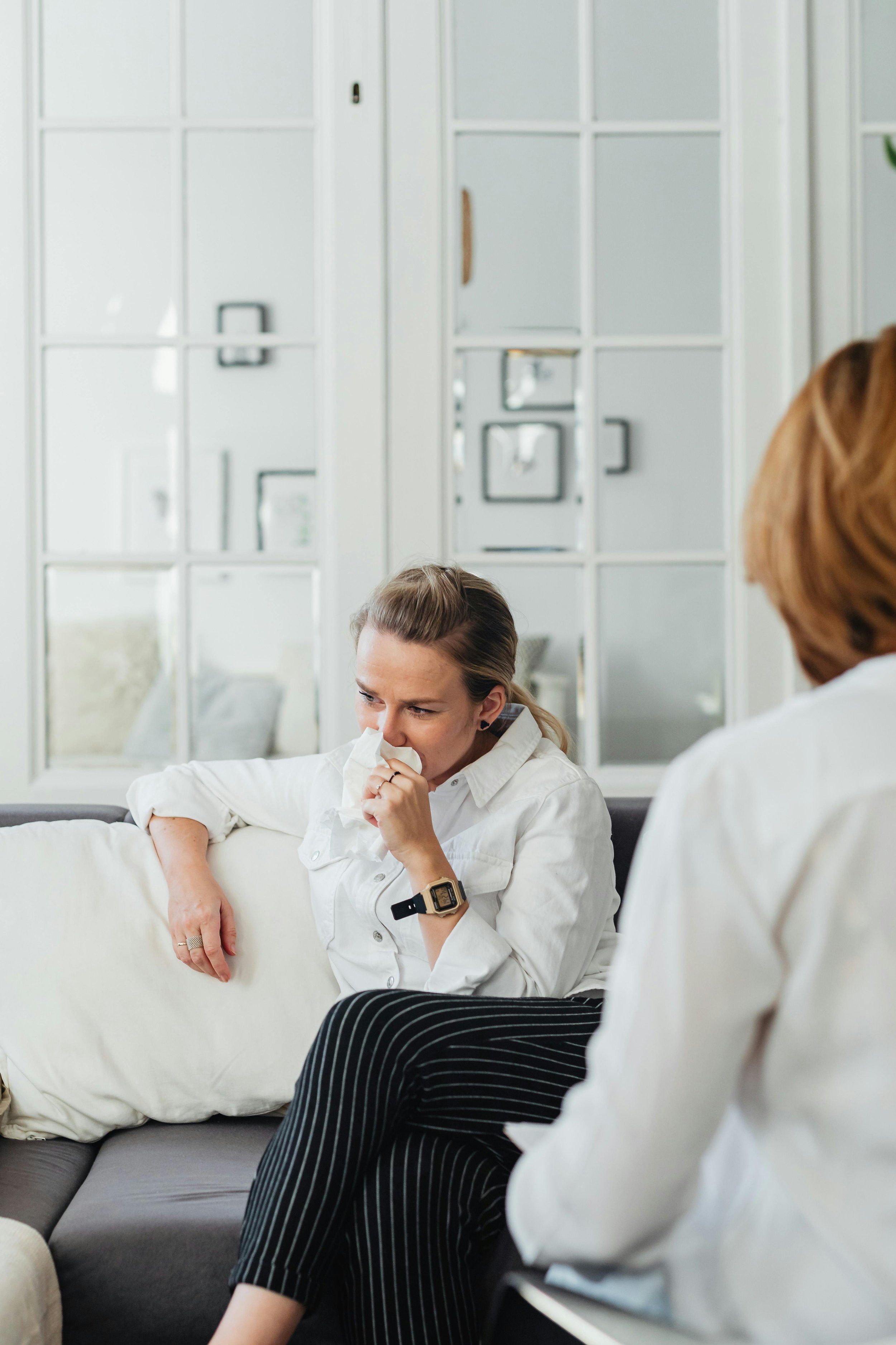 A woman sitting on a couch sneezing into a tissue, with another person sitting opposite her, in a modern living room with mirrored doors and framed pictures.