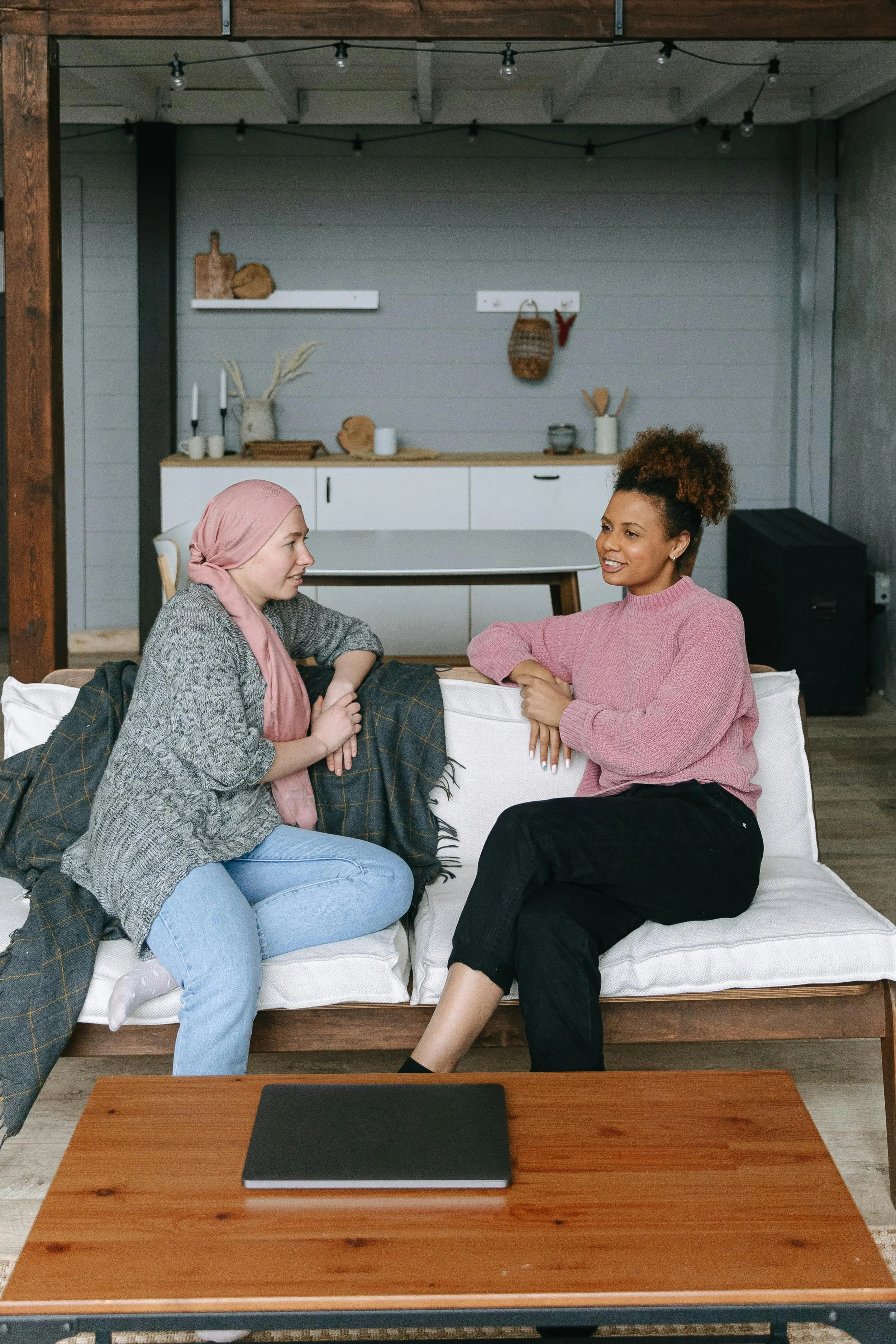 Two women having a conversation on a couch in a cozy living room, with a wooden coffee table and a laptop in front of them.