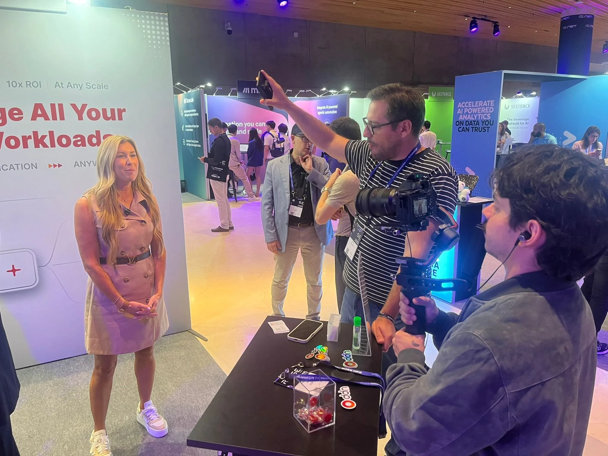 A woman being recorded by two men at a technology conference. The woman is standing near a display board, and the man is filming her with a camera mounted on a stabilizer. The other man is holding a smartphone, possibly recording or taking a picture. The scene takes place in a busy exhibition hall with various booths and attendees.