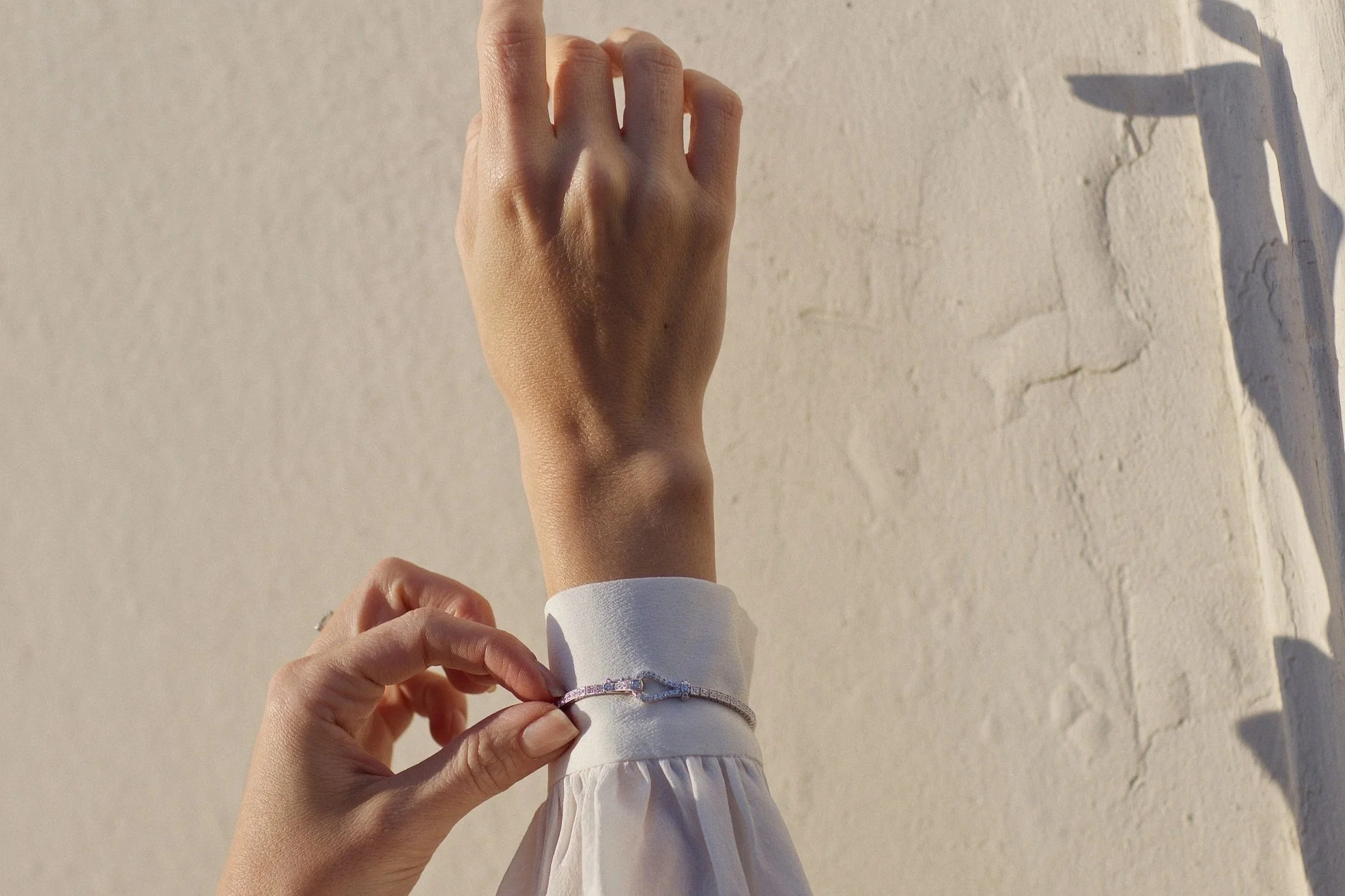 A person's hand with rings holding the sleeve of a white outfit, with a shadow of the hand cast on a textured white wall.