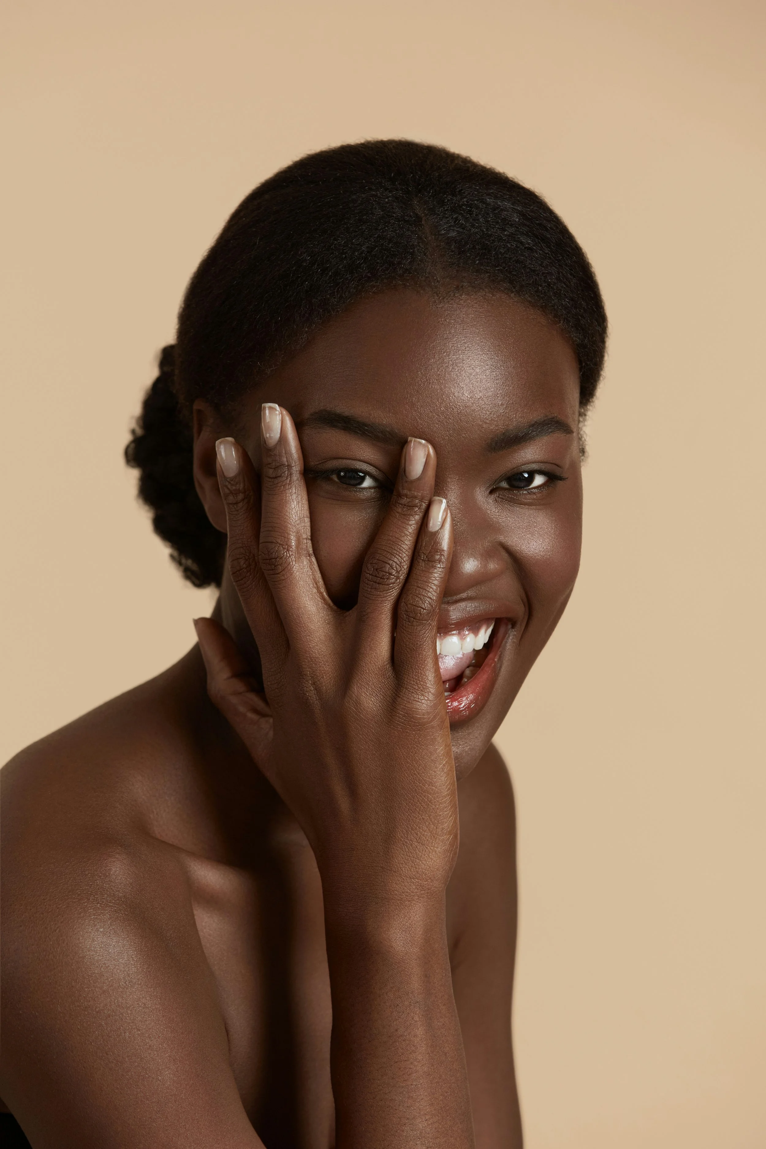 A smiling Black woman with short hair covering one eye with her hand, against a beige background.