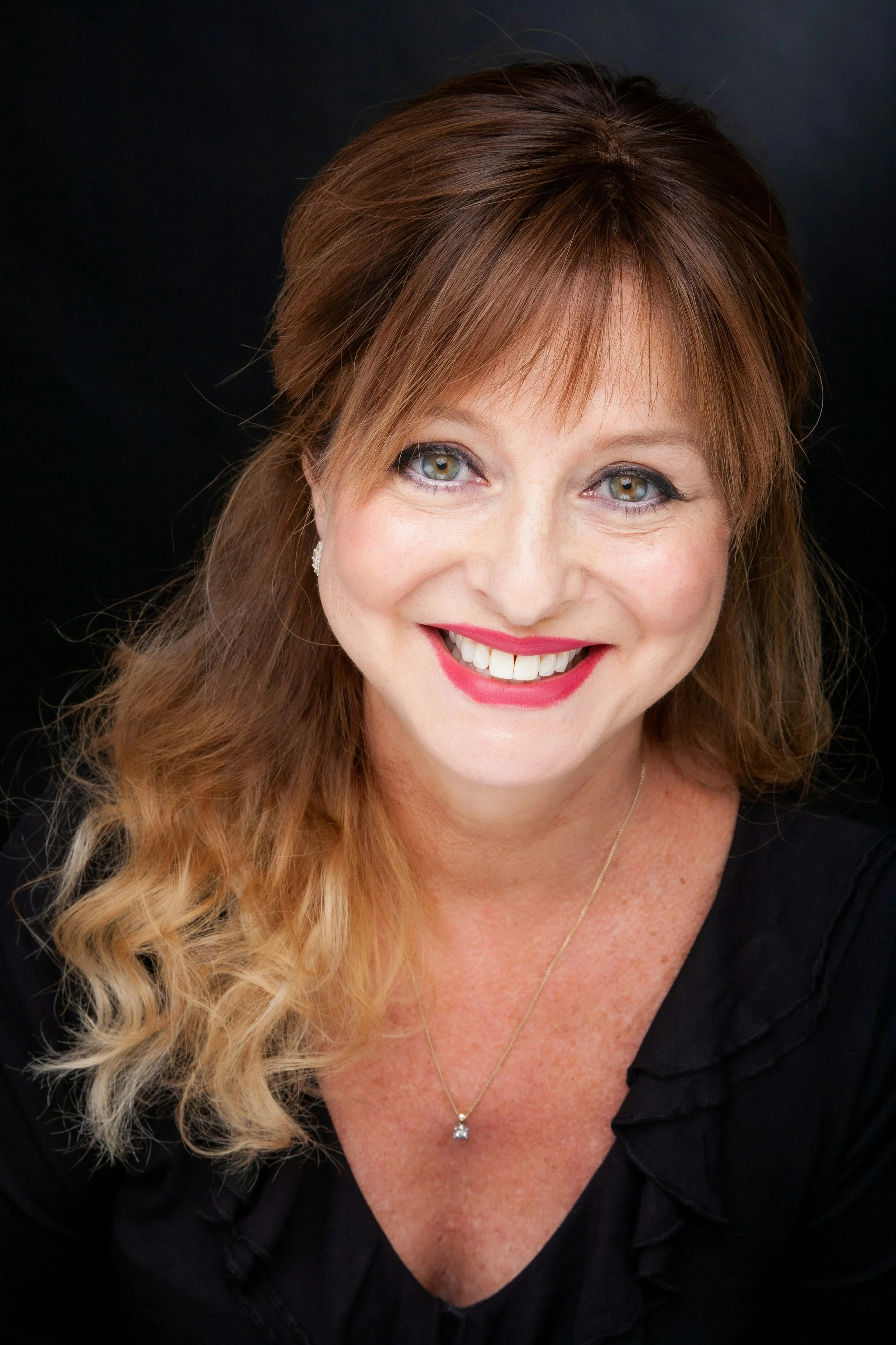 A woman with long wavy blonde hair and green eyes, smiling against a black background. She is wearing a black top, a necklace with a pendant, and earrings.
