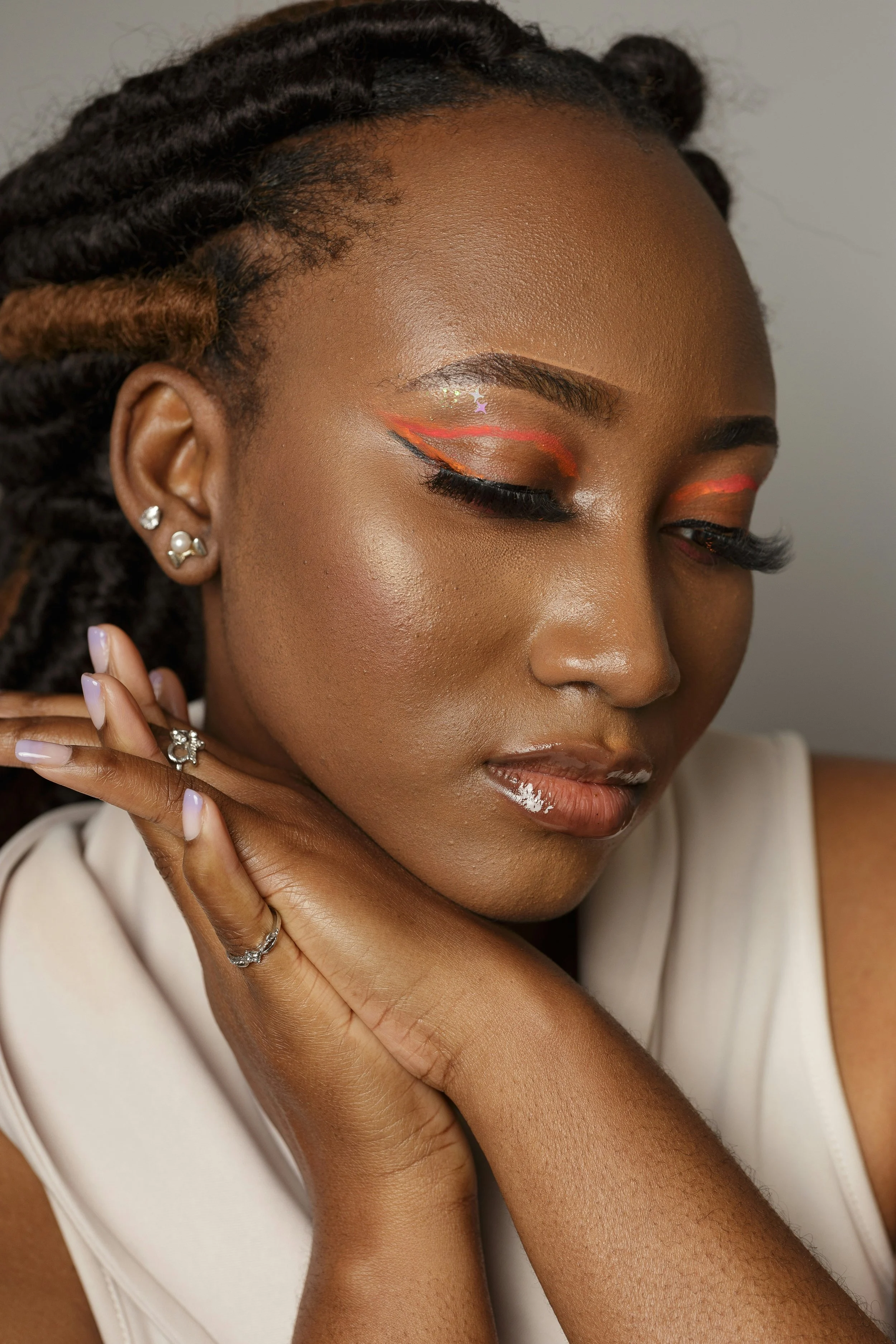 Close-up of a woman with dark skin, styled hair in twists, wearing makeup with orange and glitter eye shadow, bold eyeliner, and glossy lips, posing with her hand near her face.at Signature global Talent