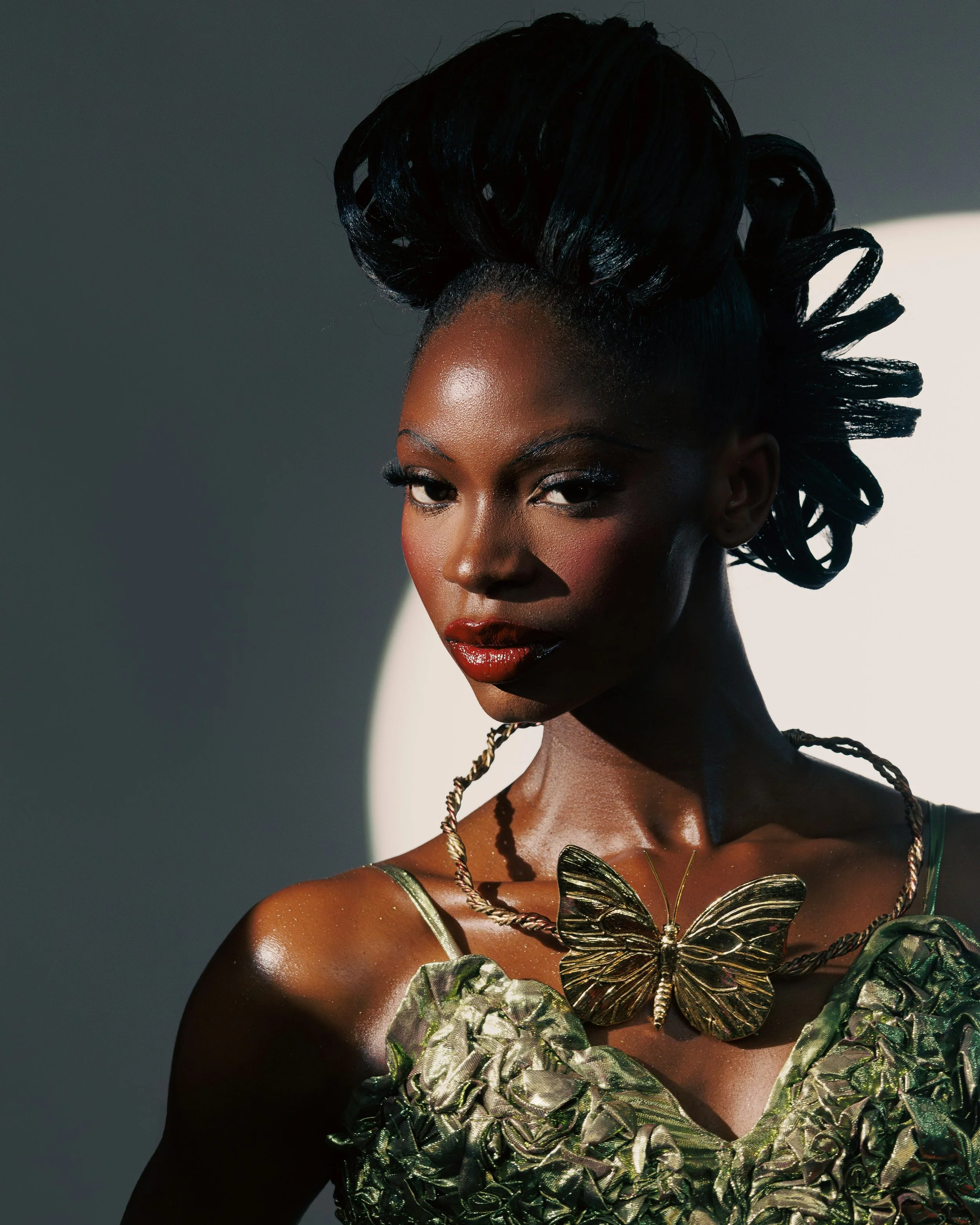 A woman with dark skin and styled black hair in an updo, wearing a gold butterfly necklace and a green textured dress, posing against a grey background.