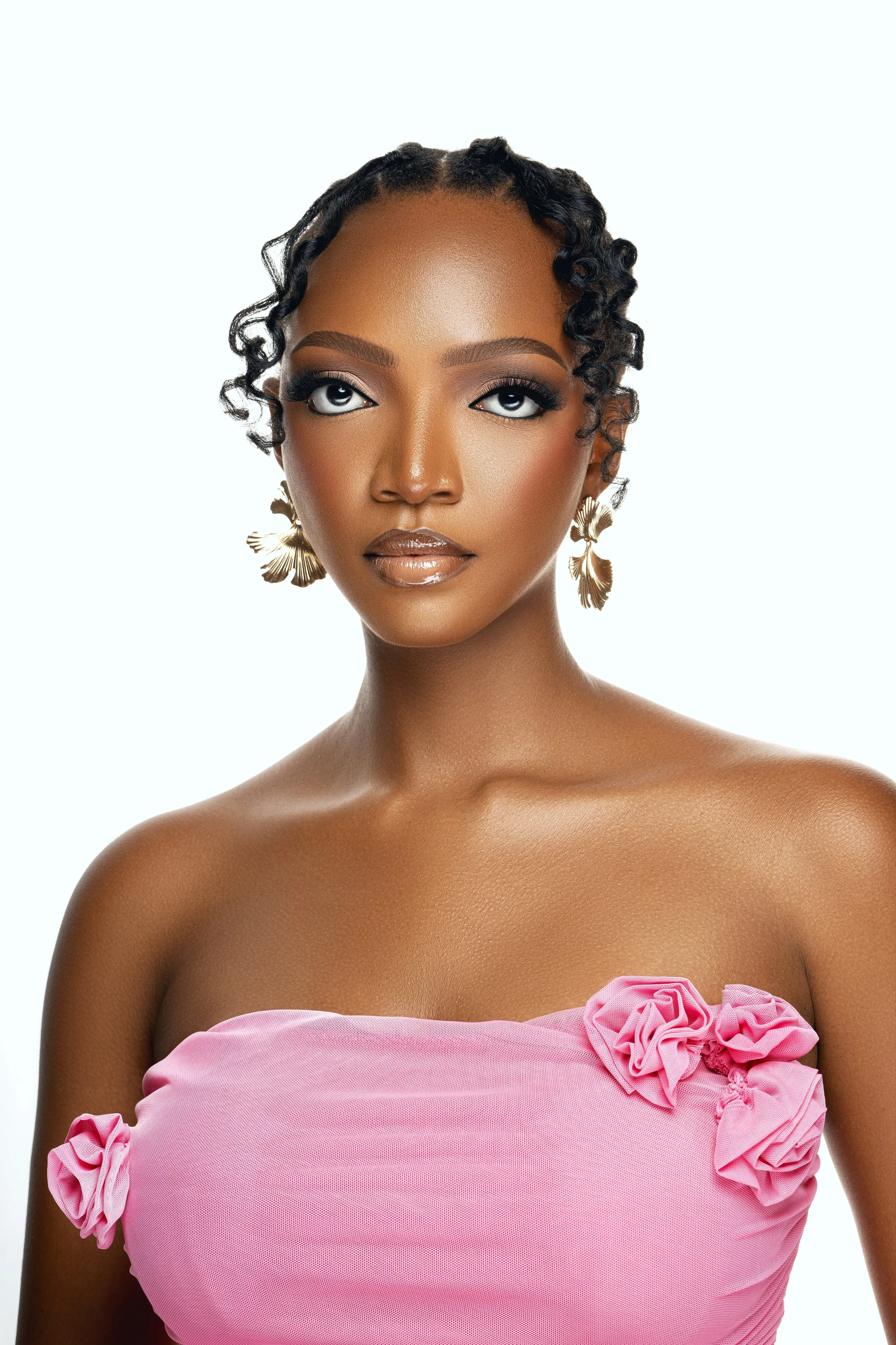 A portrait of a woman with dark skin, styled in short curly hair, wearing gold earrings, makeup, and a pink strapless top with decorative ruffles.