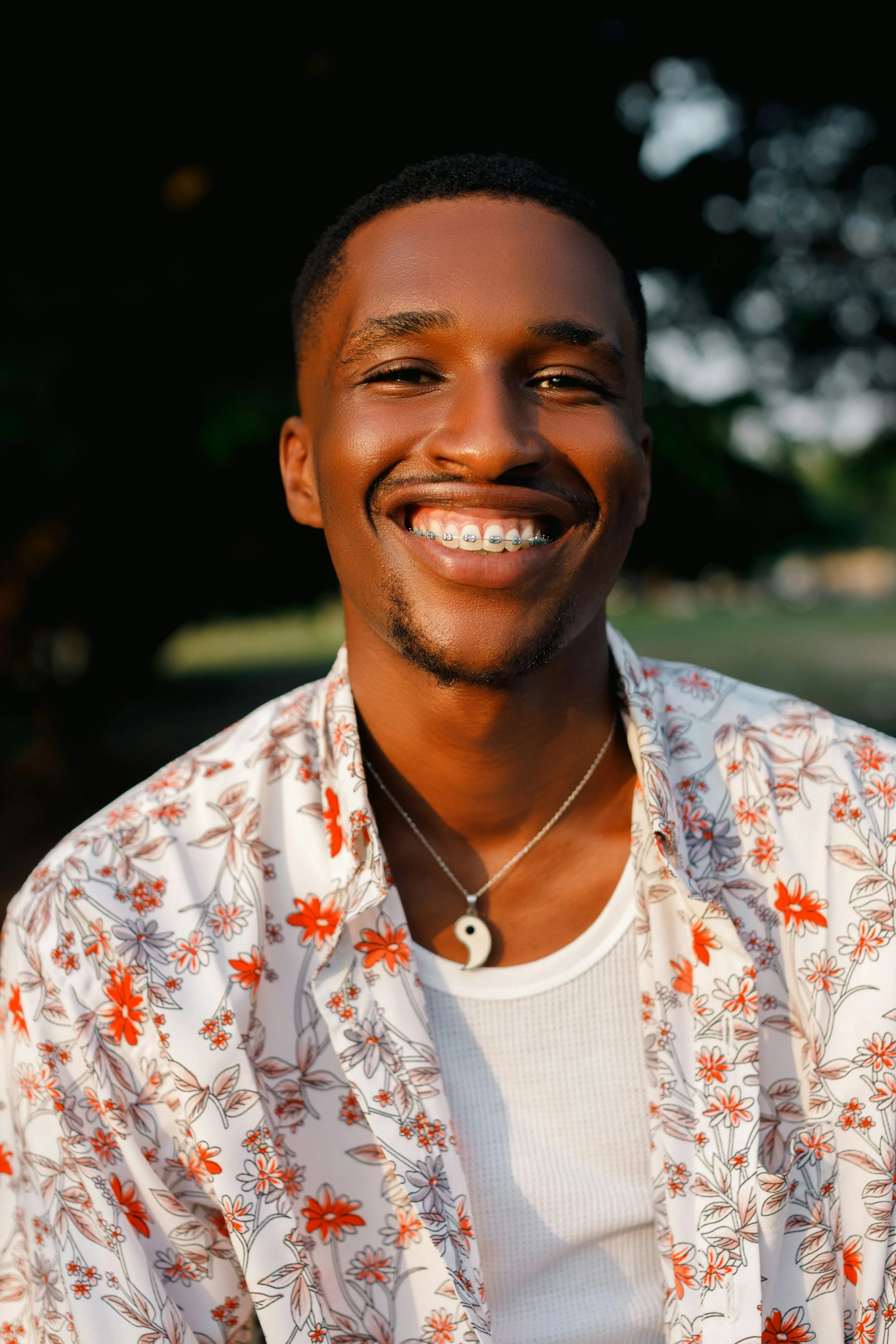 A smiling young man with braces, wearing a floral shirt, yin-yang necklace, and a white undershirt, outdoors with blurred trees in the background.