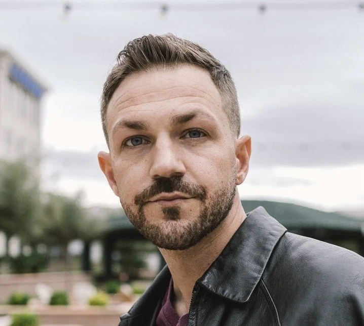 A woman with dark skin wearing a beige headwrap and large hoop earrings, posing with her head resting on her hands against a plain white background.