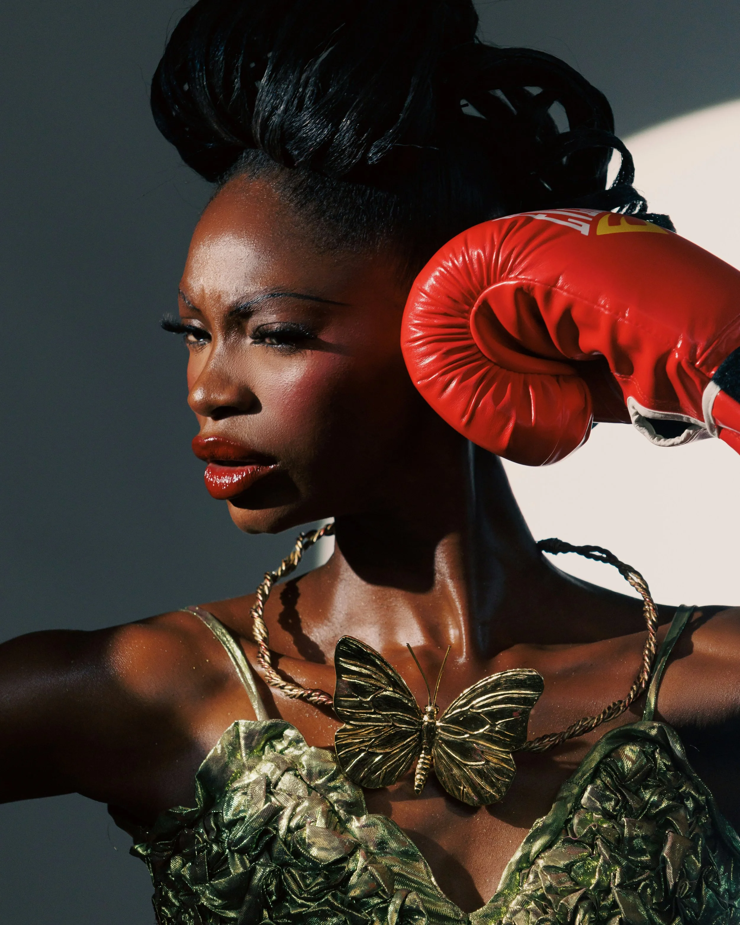 A woman with styled hair wears a green dress, gold butterfly necklace, and red boxing glove on one hand, posing with a serious expression.at Signature global Talent