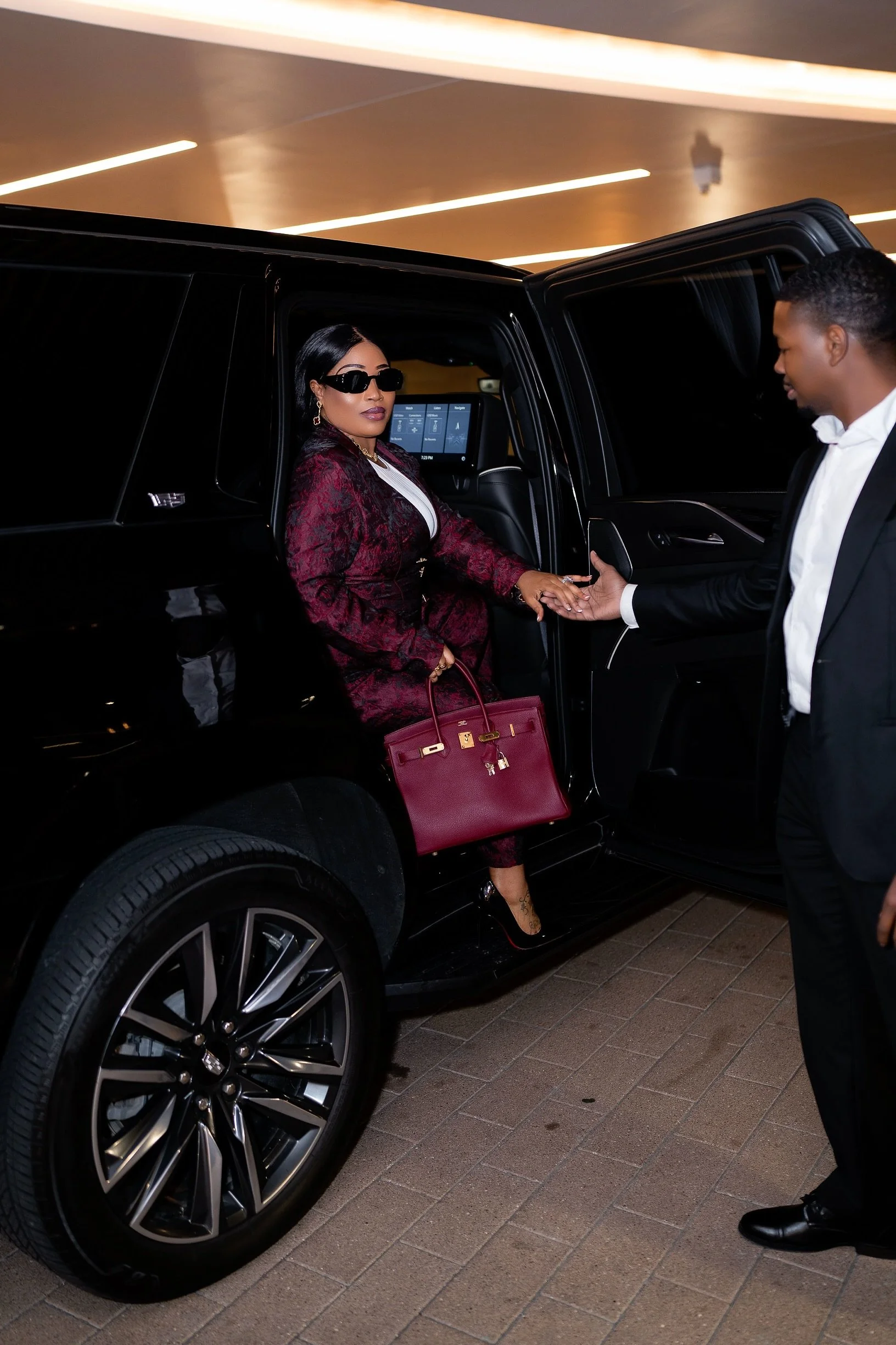 A woman dressed in a burgundy outfit with a matching handbag, sunglasses, and high heels is getting out of a black luxury SUV while a man in a black tuxedo assists her.