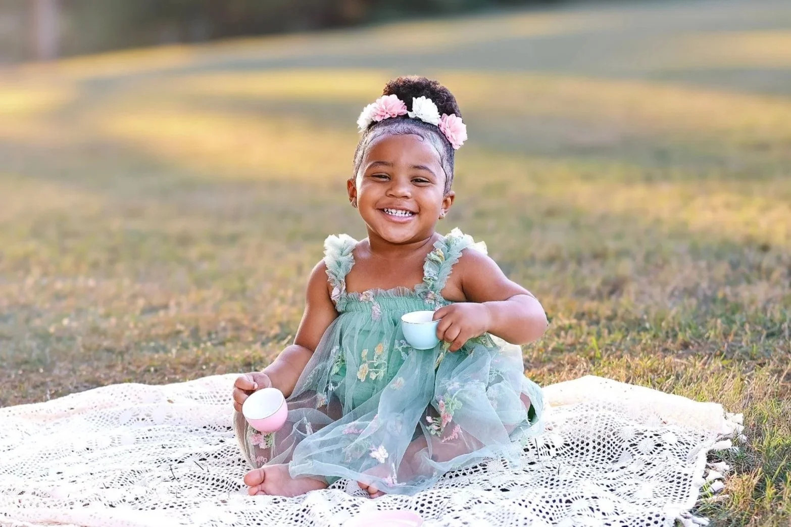 A happy young girl with a floral headband and a green dress sitting on a white blanket outdoors, holding a teacup, smiling at the camera.