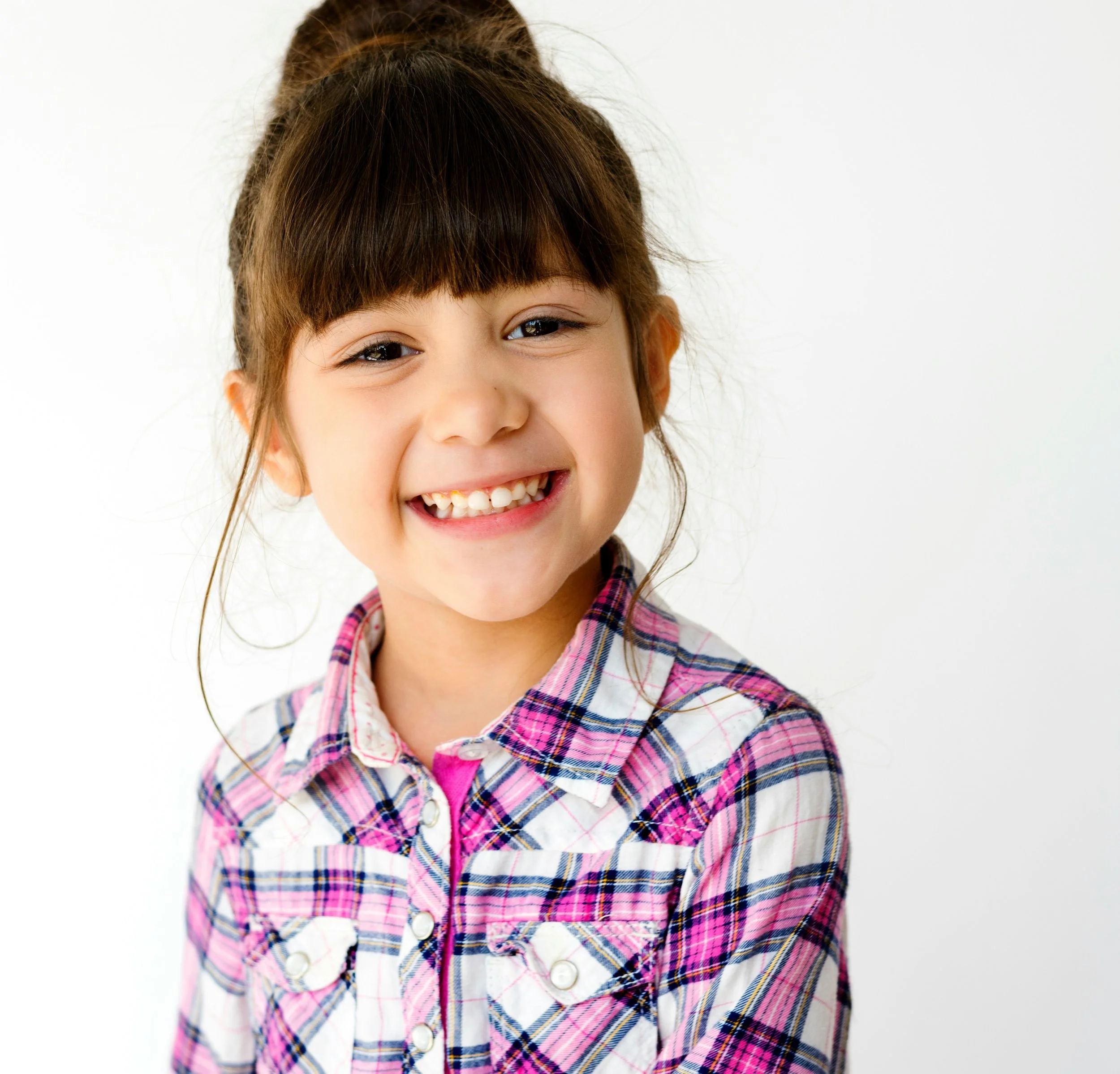 A young girl smiling at the camera, with brown hair in a ponytail, wearing a pink, purple, and white plaid shirt against a white background.