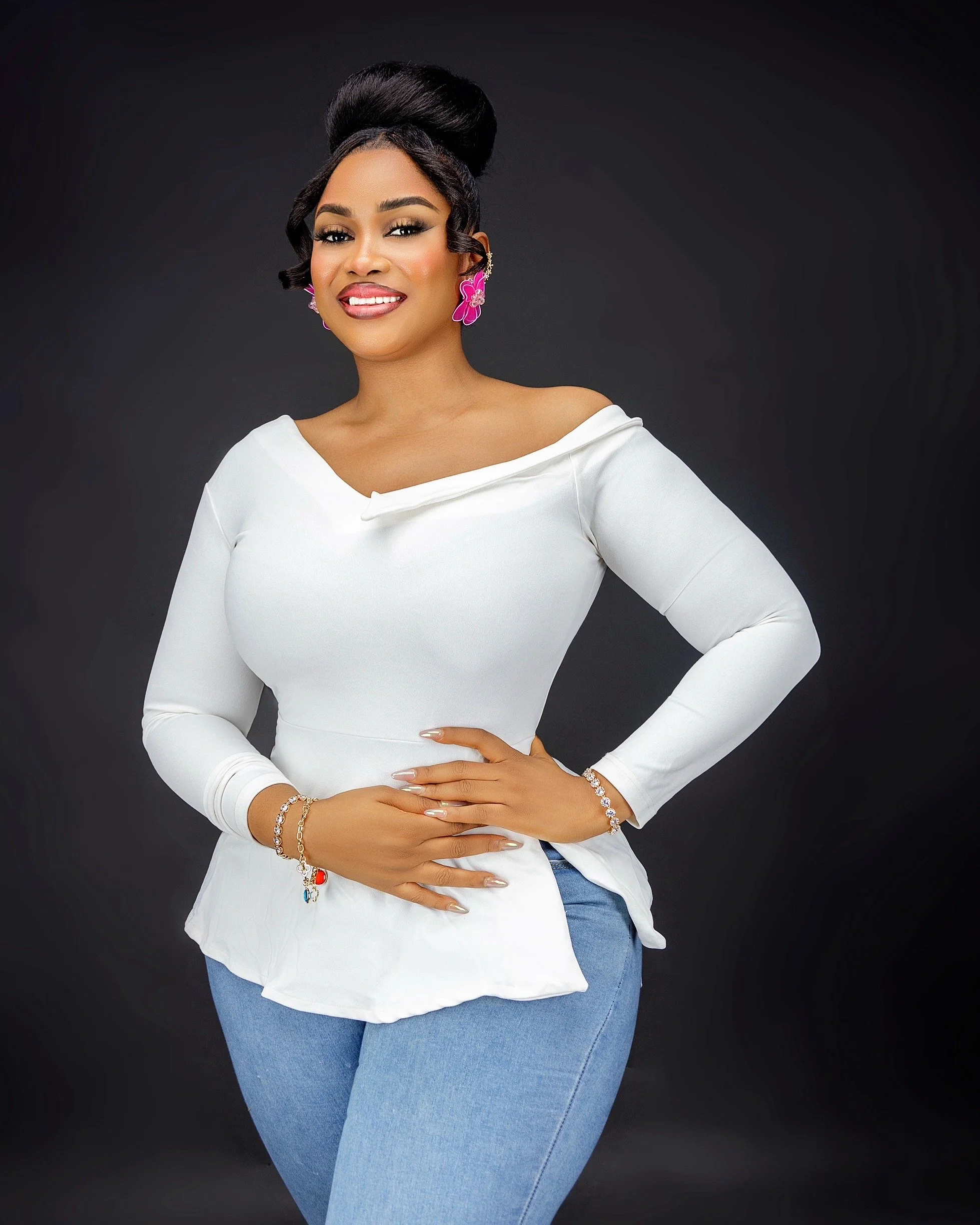 A woman with styled hair, wearing a white top and jeans, standing against a dark background with her hand on her stomach, smiling.at Signature global Talent