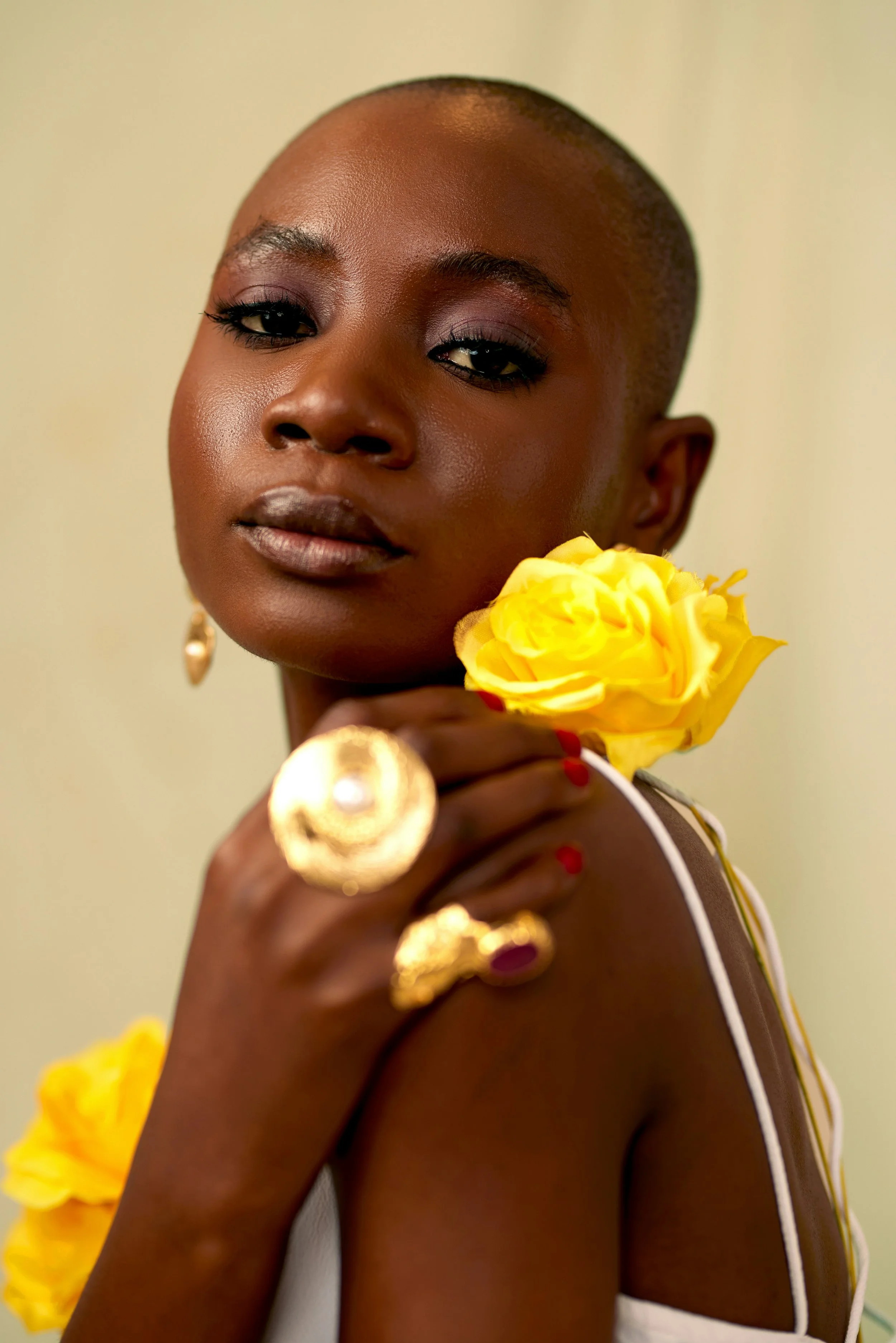 A close-up portrait of a woman with dark skin, short hairstyle, and elegant makeup. She is holding a yellow flower near her face and wearing large gold rings and earrings. The background is plain and light-colored.