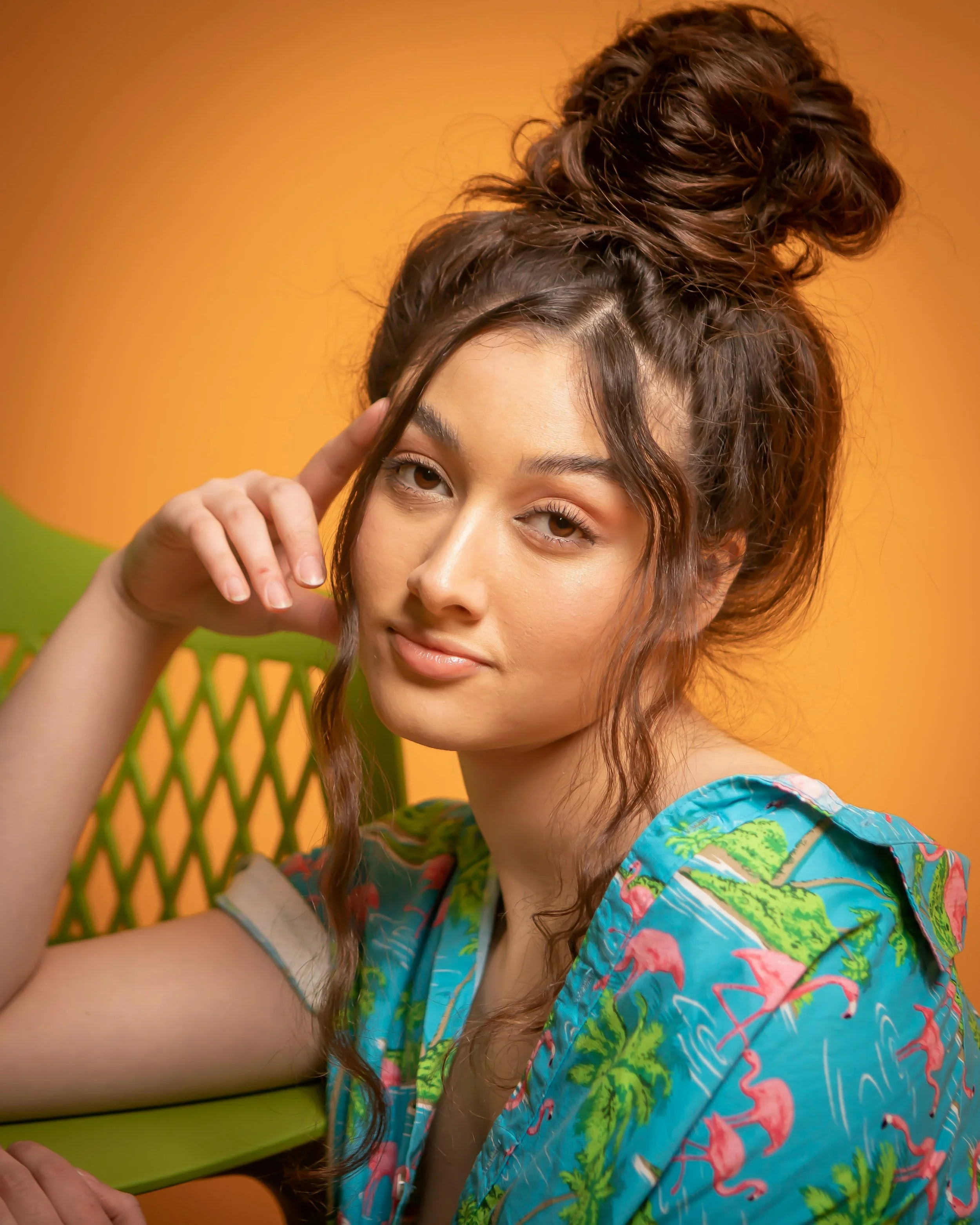 A young woman with wavy brown hair styled in a high messy bun, wearing a colorful tropical shirt, sitting against an orange background with her elbow resting on a green chair, looking at the camera with a relaxed expression.
