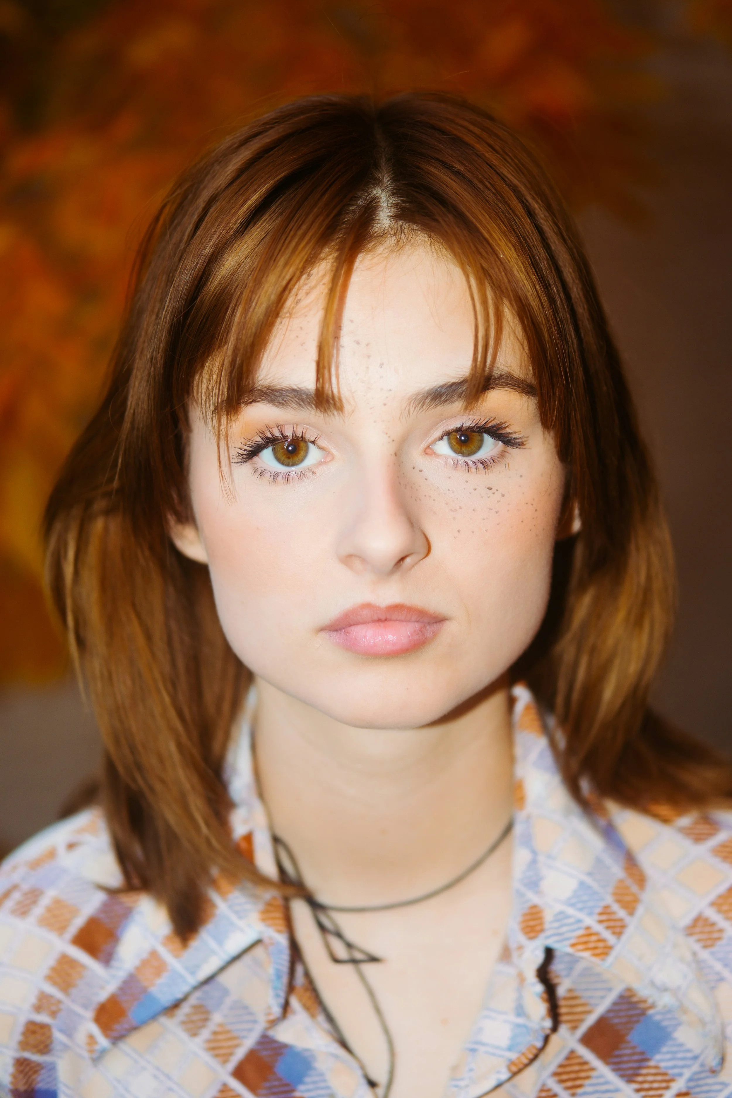Close-up portrait of a young woman with reddish-brown hair, hazel eyes, and freckles, wearing a plaid shirt and a black choker necklace.