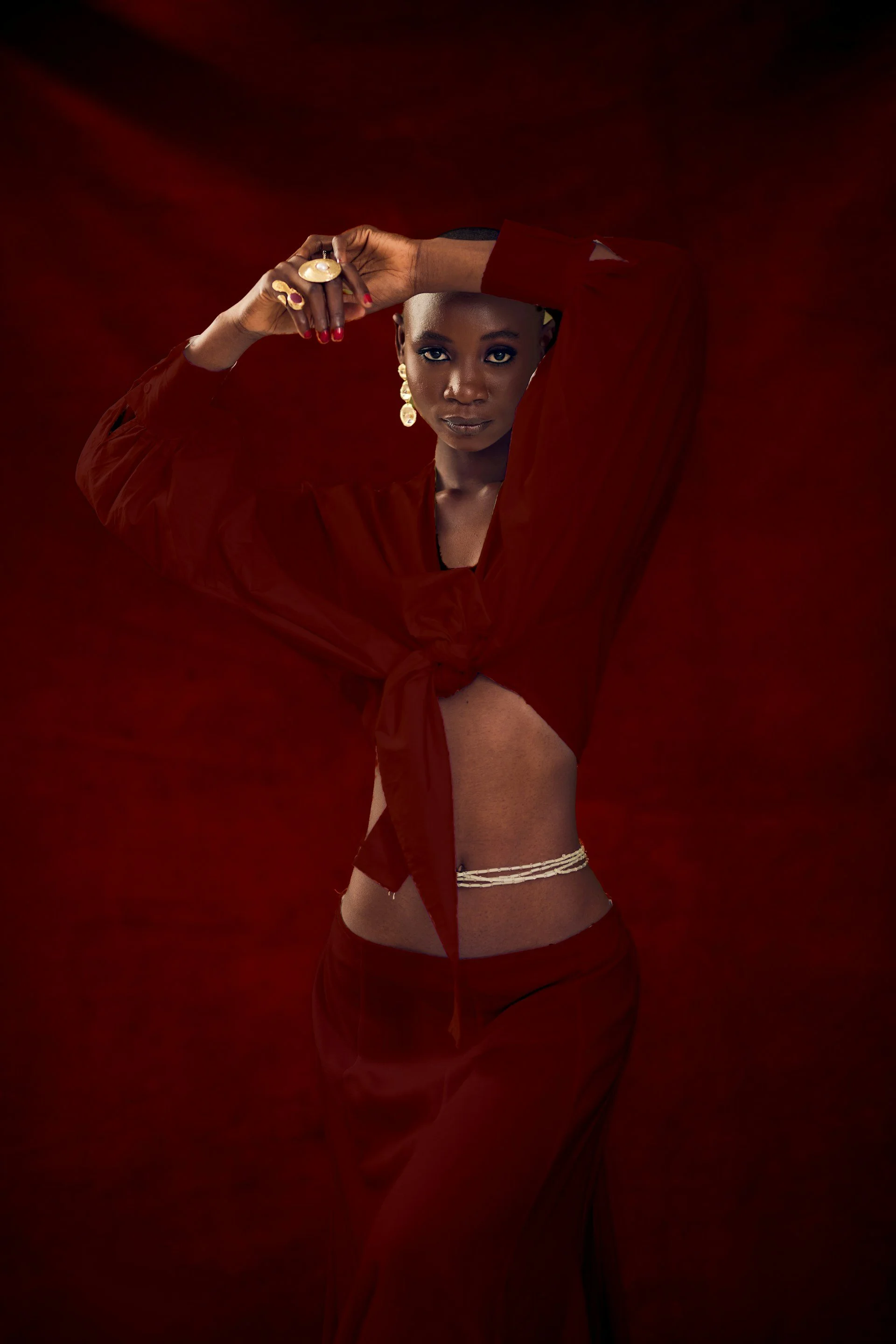 A woman with dark skin and jewelry poses against a dark red background, wearing a red long-sleeved top tied at her waist, revealing her midriff and accessories.
