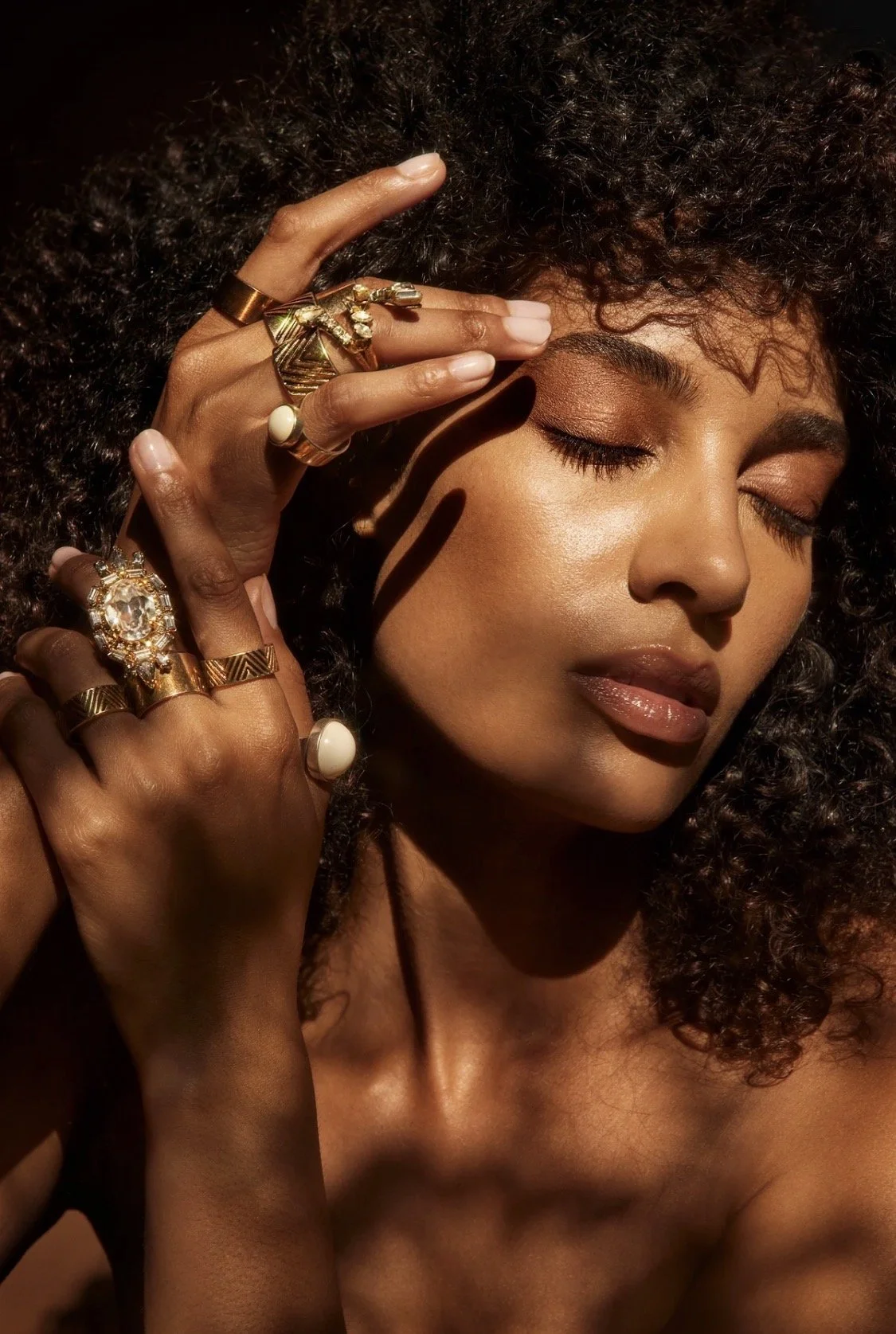 A woman with dark curly hair closed her eyes, showcasing gold jewelry and rings on her fingers, with a serene expression on her face.at Signature global Talent