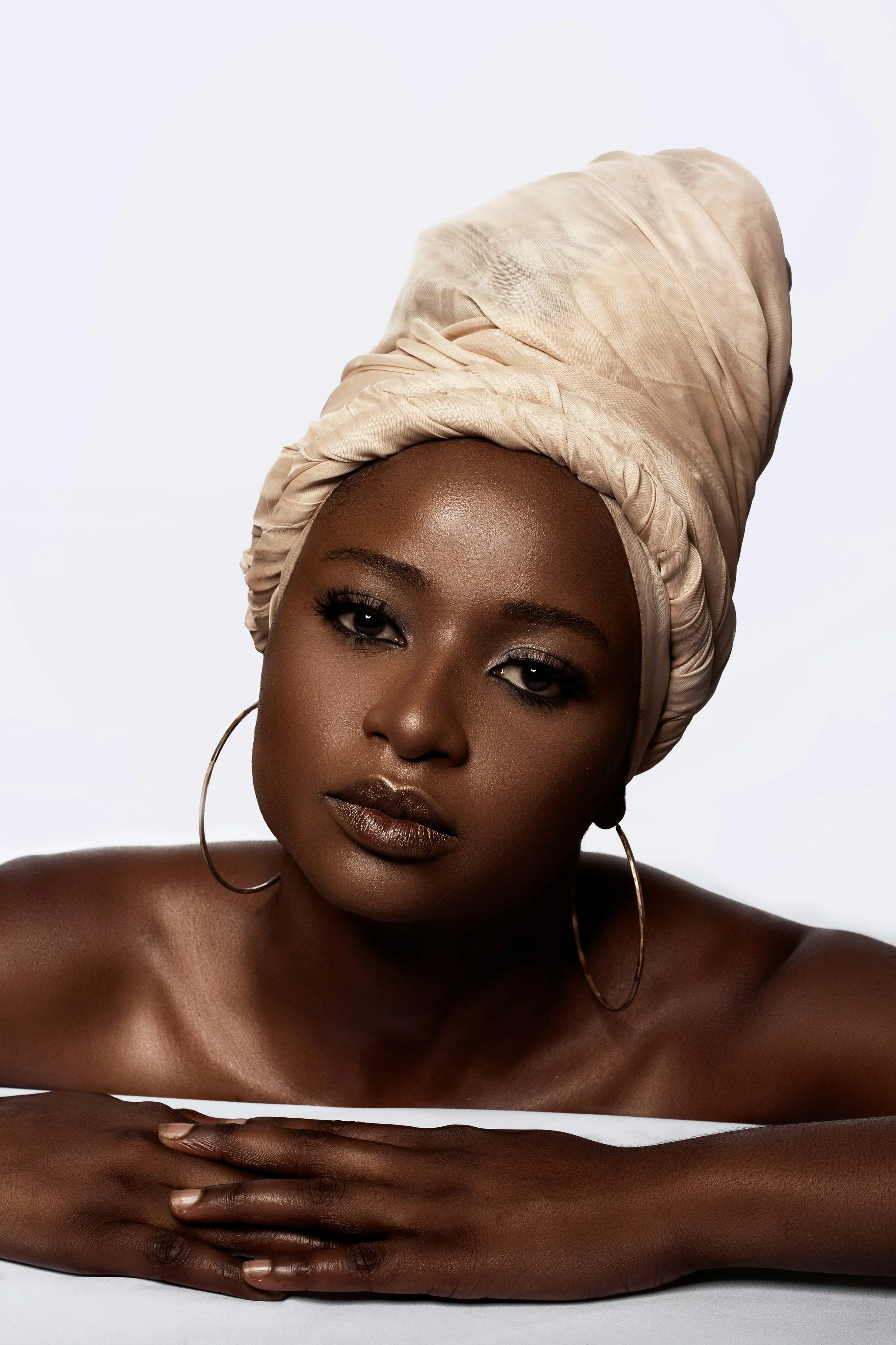 A woman with dark skin wearing a beige headwrap and large hoop earrings, posing with her head resting on her hands against a plain white background.