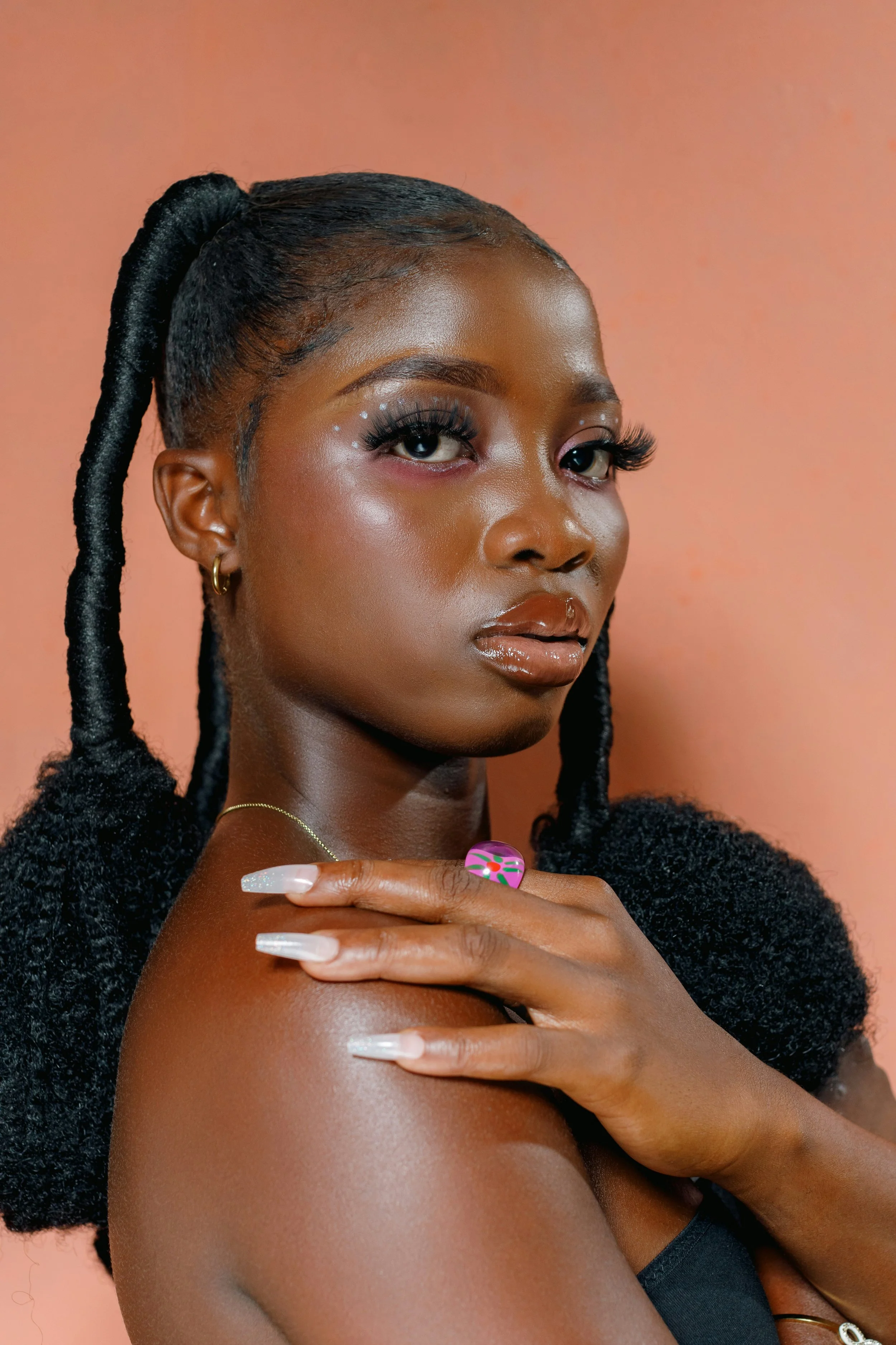 A young woman with dark skin and long braided hair styled in a high ponytail. She is wearing makeup with long eyelashes and shimmering eyeshadow, and has glossy lips. Her hand with long, manicured nails rests on her shoulder, and she is wearing a black top, gold hoop earrings, a delicate gold necklace, and a colorful ring. The background is a soft peach color.