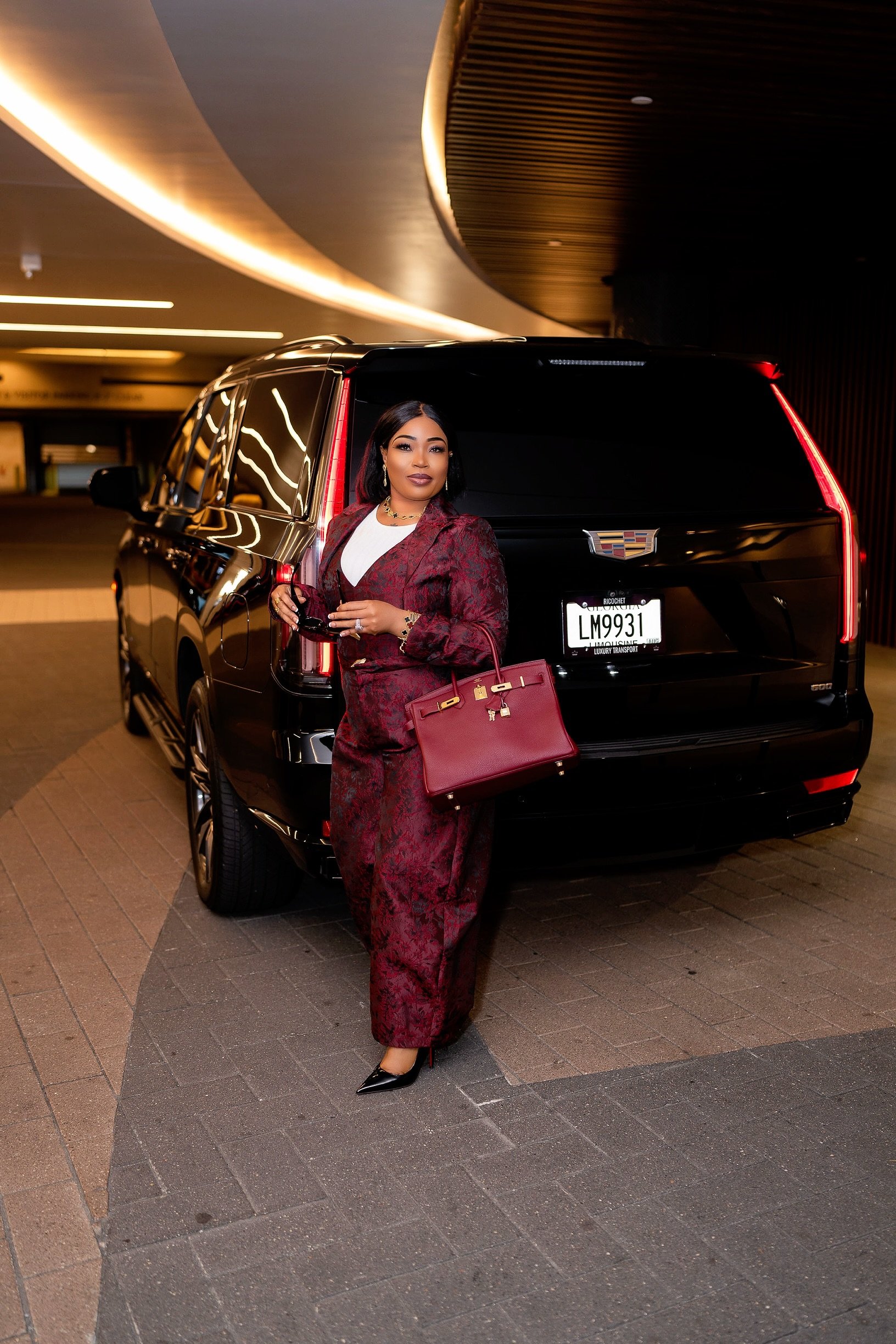 A woman dressed in a red patterned suit with a white top, standing next to a black luxury Cadillac SUV in an indoor parking garage, holding a red handbag and wearing black high heels.