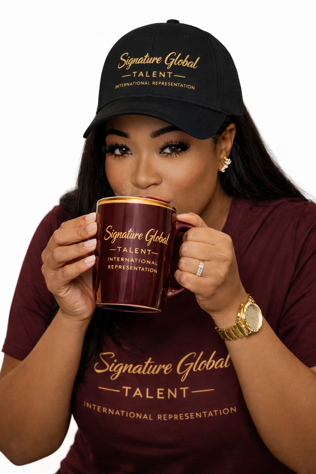A woman wearing a black cap and maroon T-shirt with gold text, holding a large maroon mug with gold writing, sipping from it. She has long dark hair, gold earrings, a ring, a gold watch, and is looking at the camera.at Signature global Talent