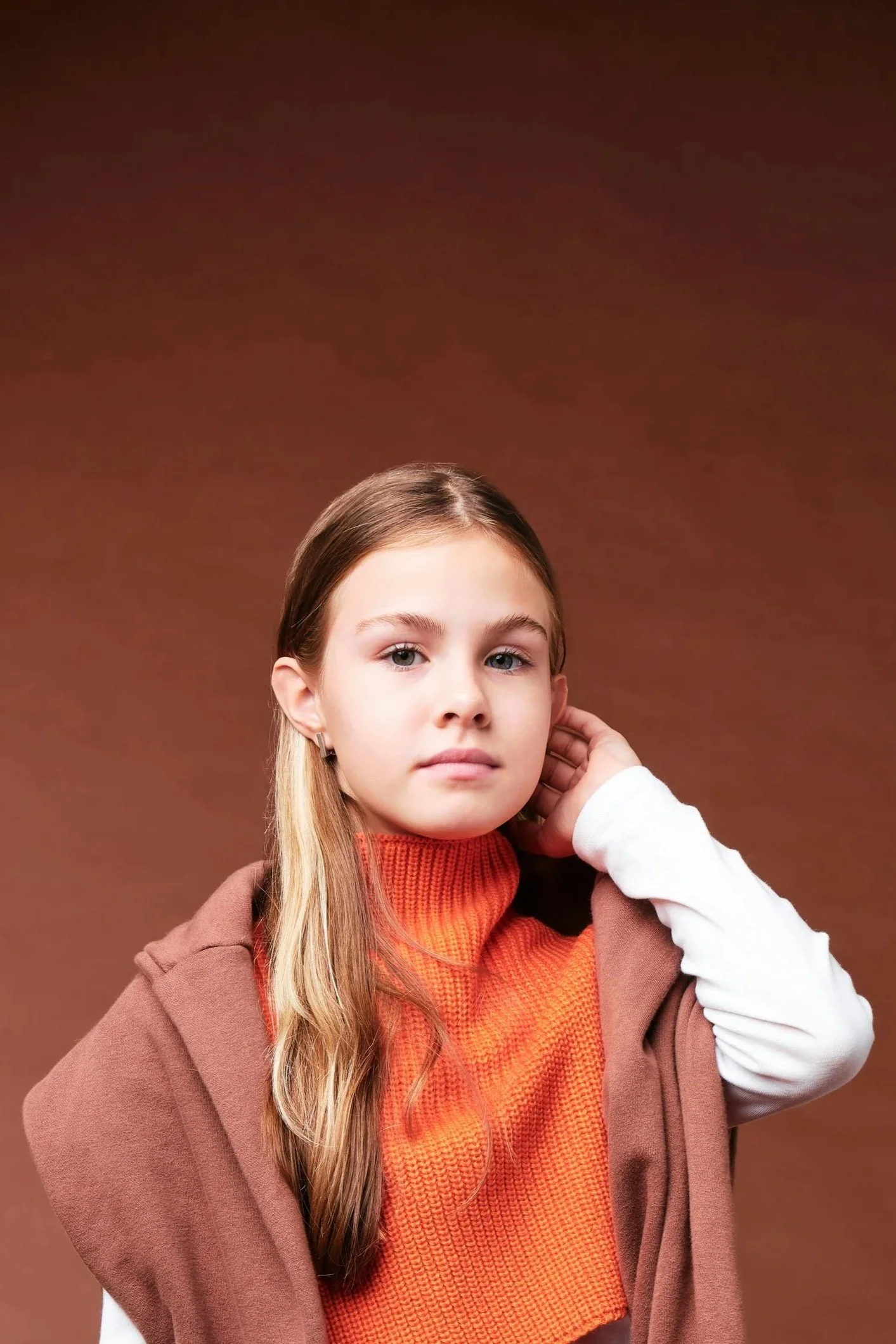 Portrait of a young girl with long blonde hair wearing an orange turtleneck sweater and a brown coat, standing against a brown background.