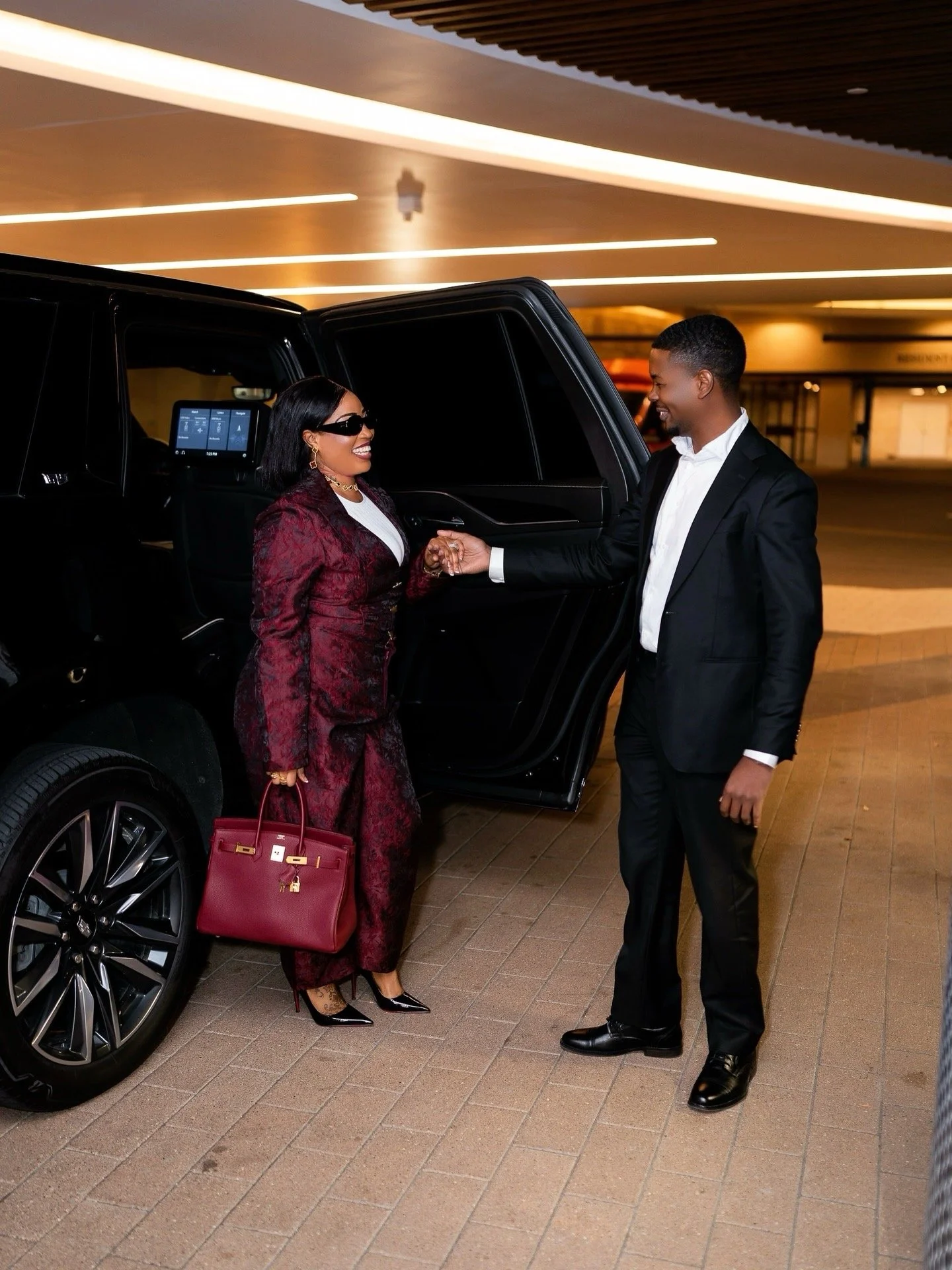 A woman in a maroon suit and sunglasses is smiling and shaking hands with a man in a black suit outside a black luxury vehicle in a parking garage.