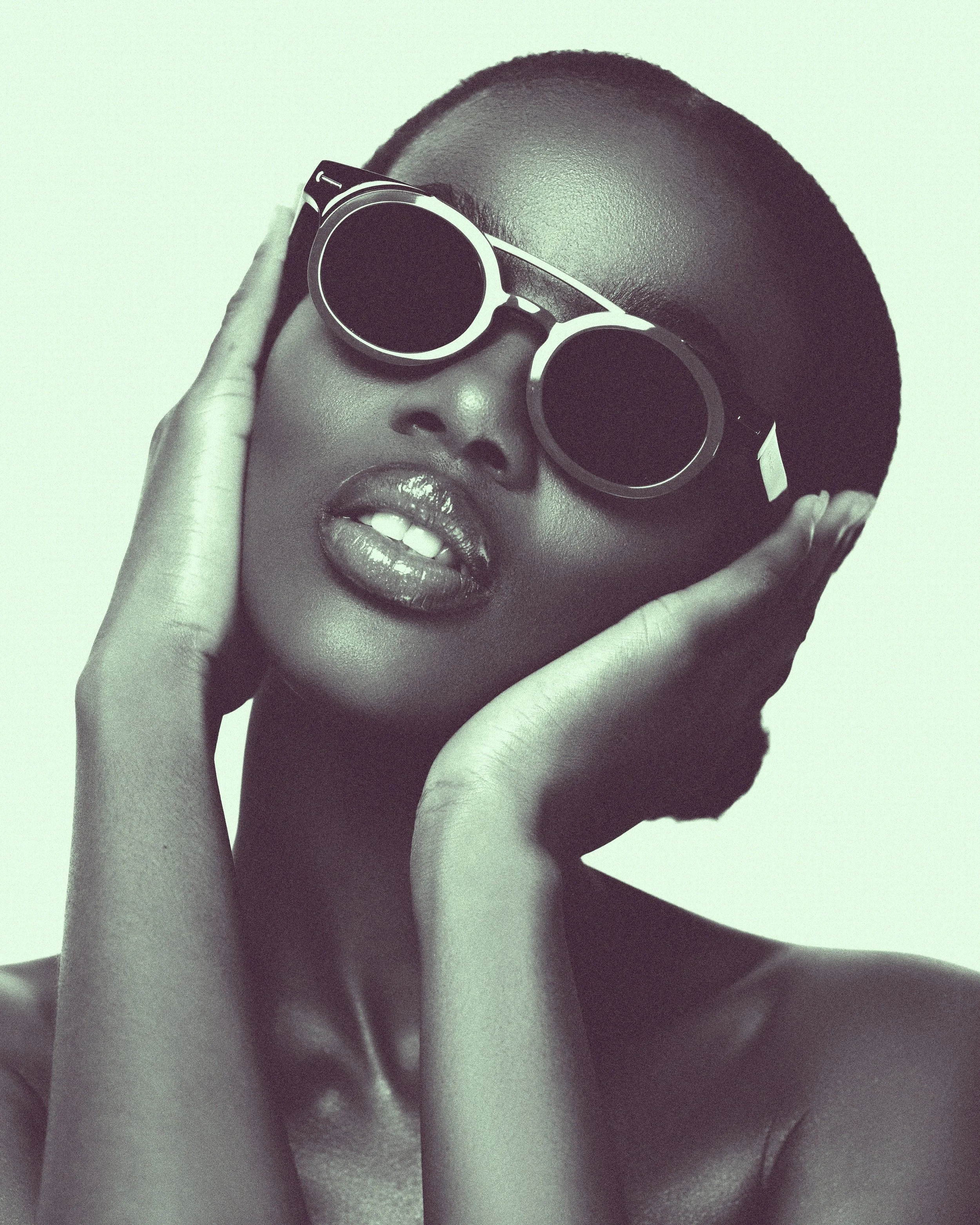 A woman with short hair wearing dark sunglasses and shiny lipstick, touching her face with her hands.