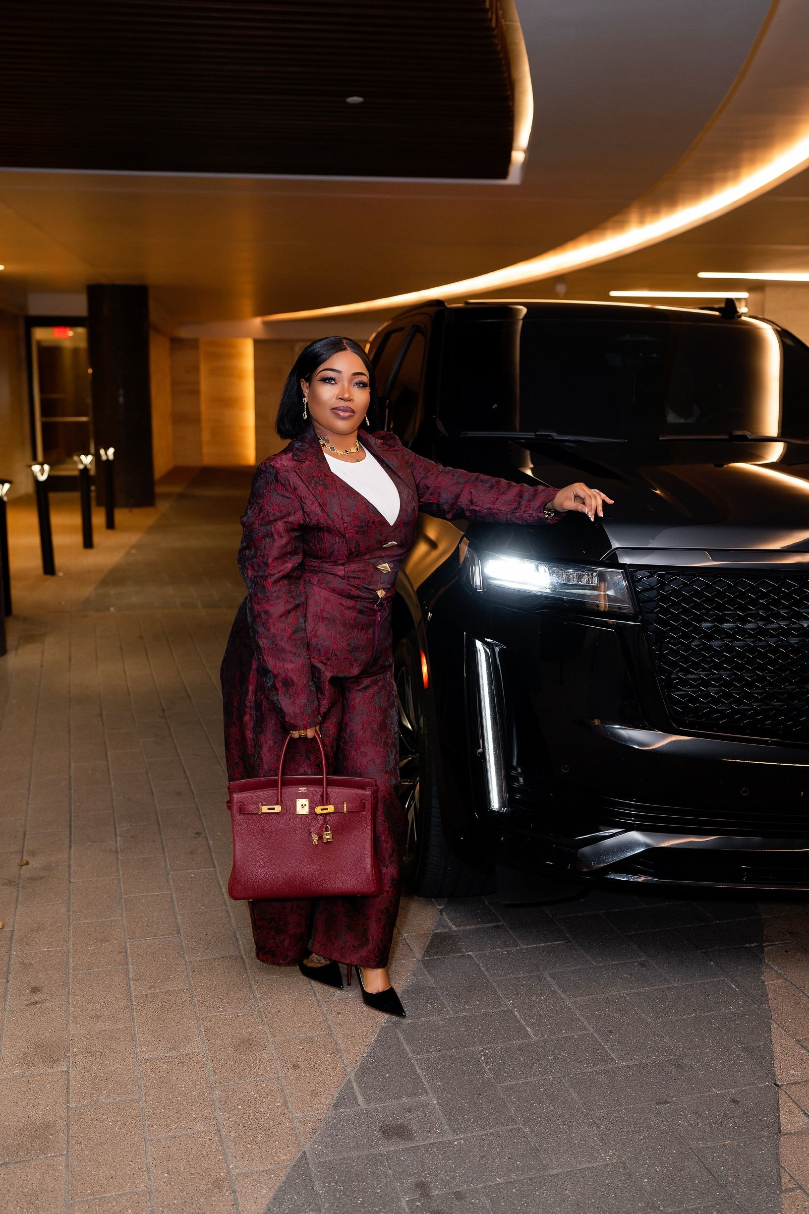 A woman dressed in a maroon patterned suit holding a matching maroon handbag stands next to a black SUV in a modern parking garage with warm lighting.