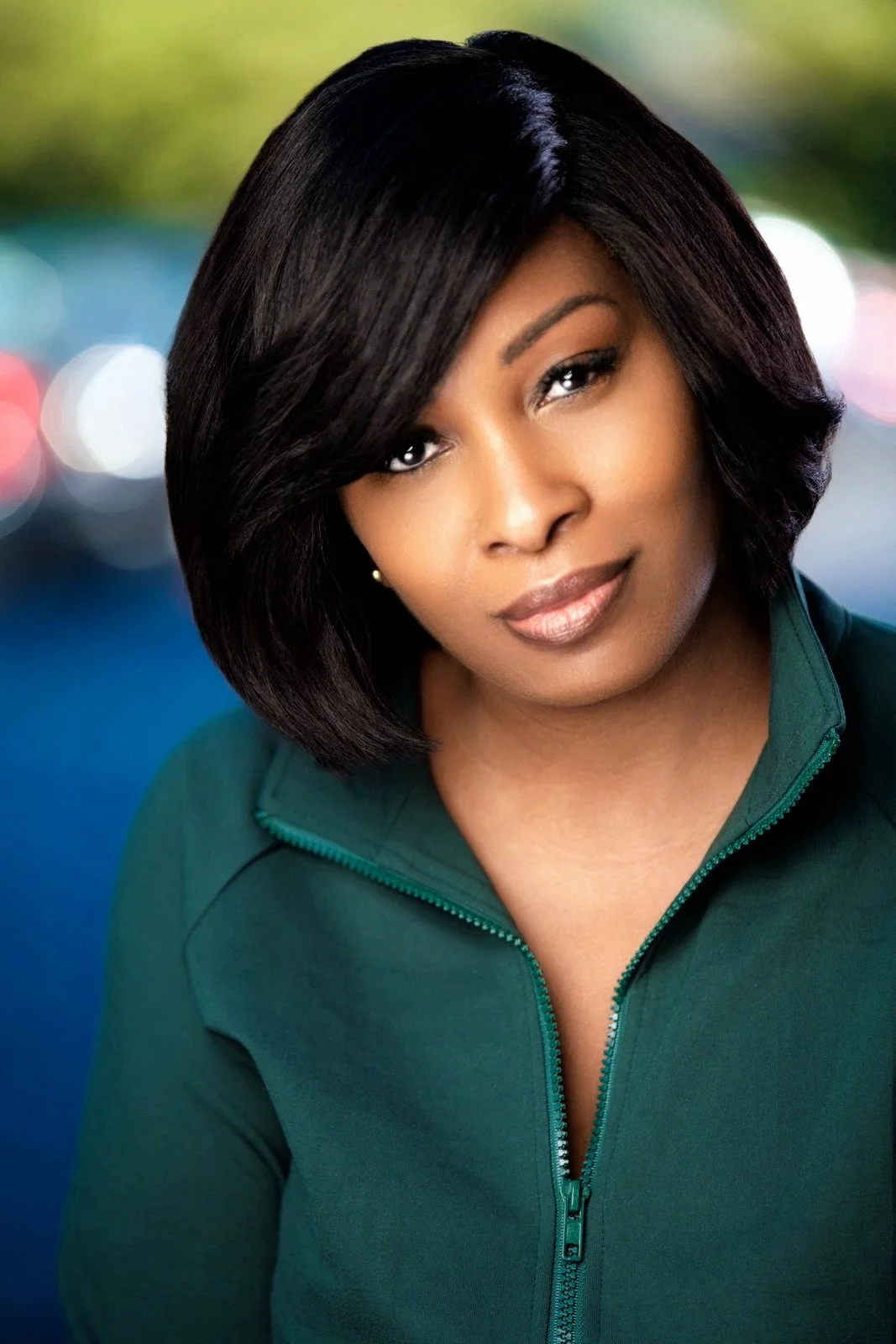 A woman with shoulder-length black hair and brown skin, wearing a dark green zip-up jacket, looking at the camera with a slight smile, blurred colorful lights in the background. at Signature global Talent