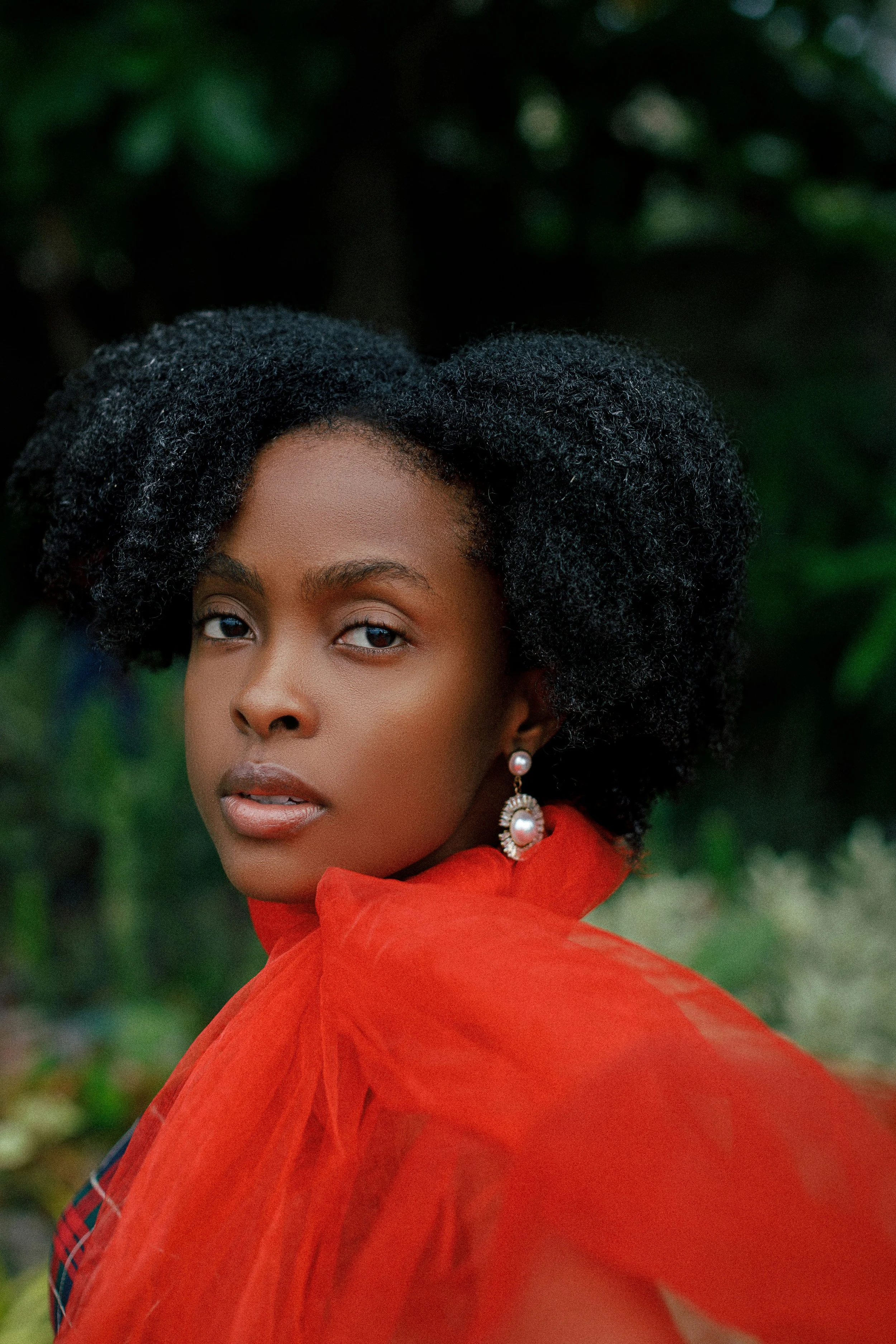 A young African American woman with natural curly hair, wearing large pearl earrings and a red sheer garment, is outdoors with a blurred green foliage background.