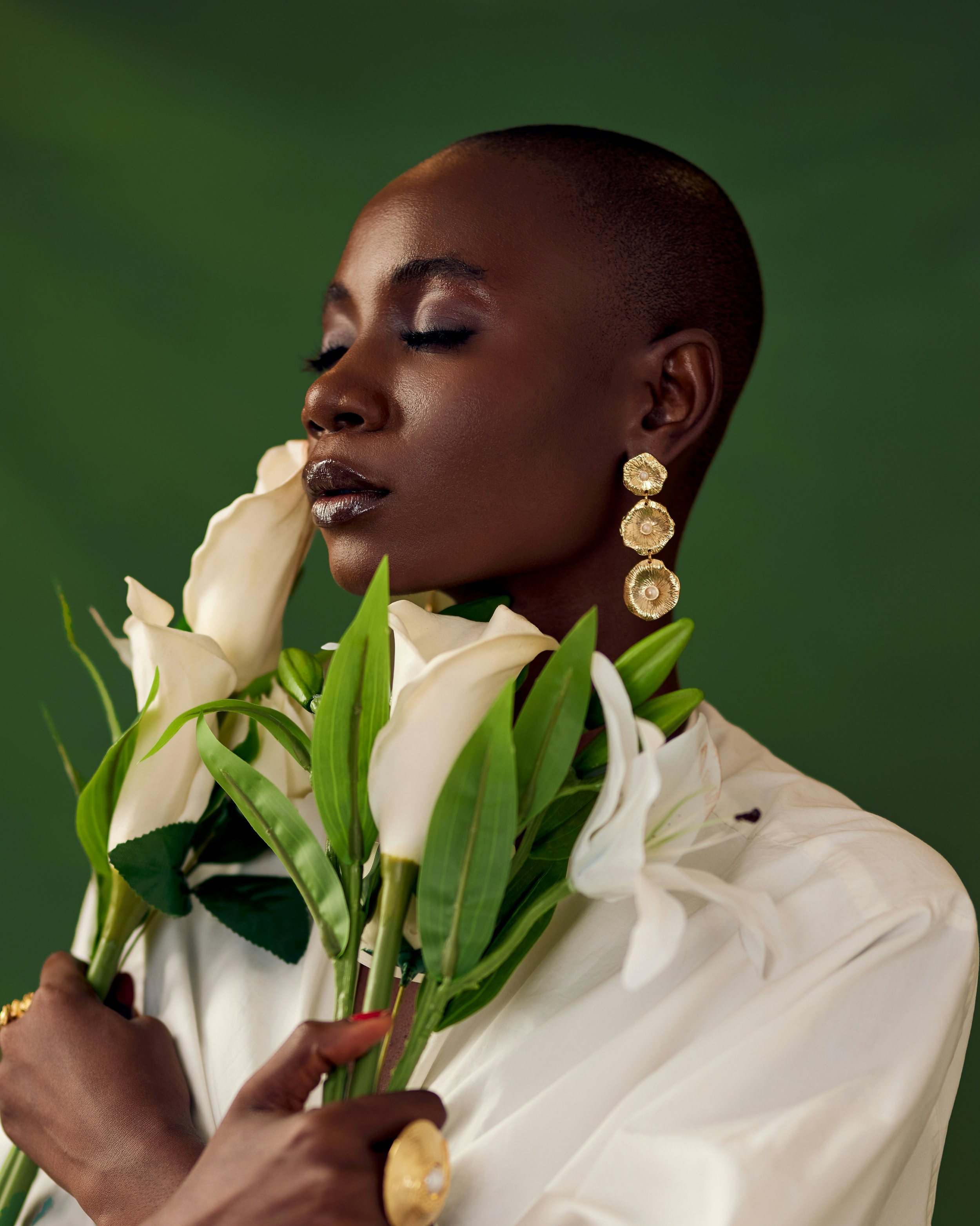 A woman with dark skin and a shaved head holding white calla lilies and wearing large gold earrings and a cream-colored blouse against a green background.