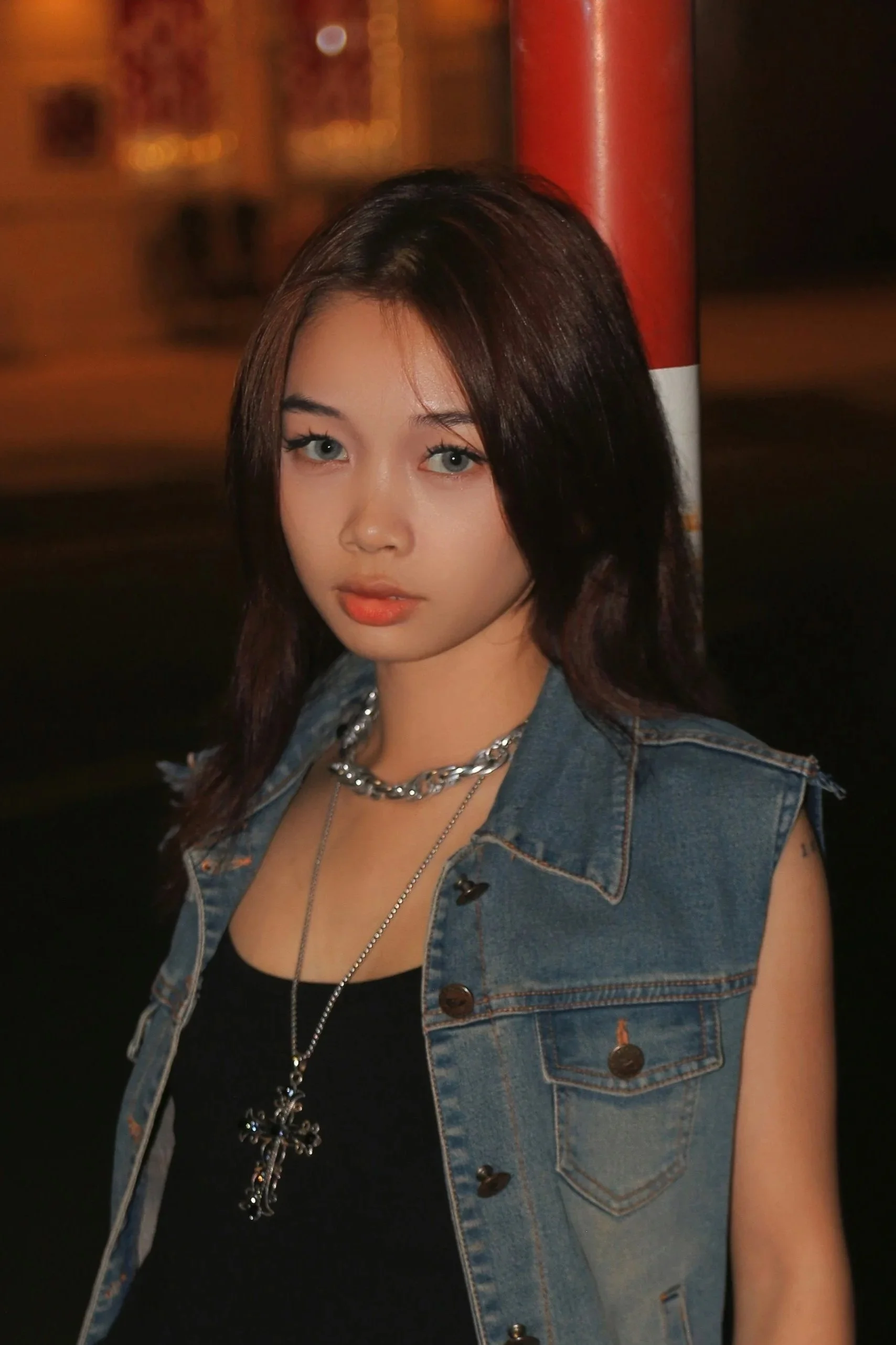 A young woman with dark hair and blue eyes stands outdoors at night, wearing a sleeveless denim vest over a black top, accessorized with layered silver necklaces including a cross pendant.