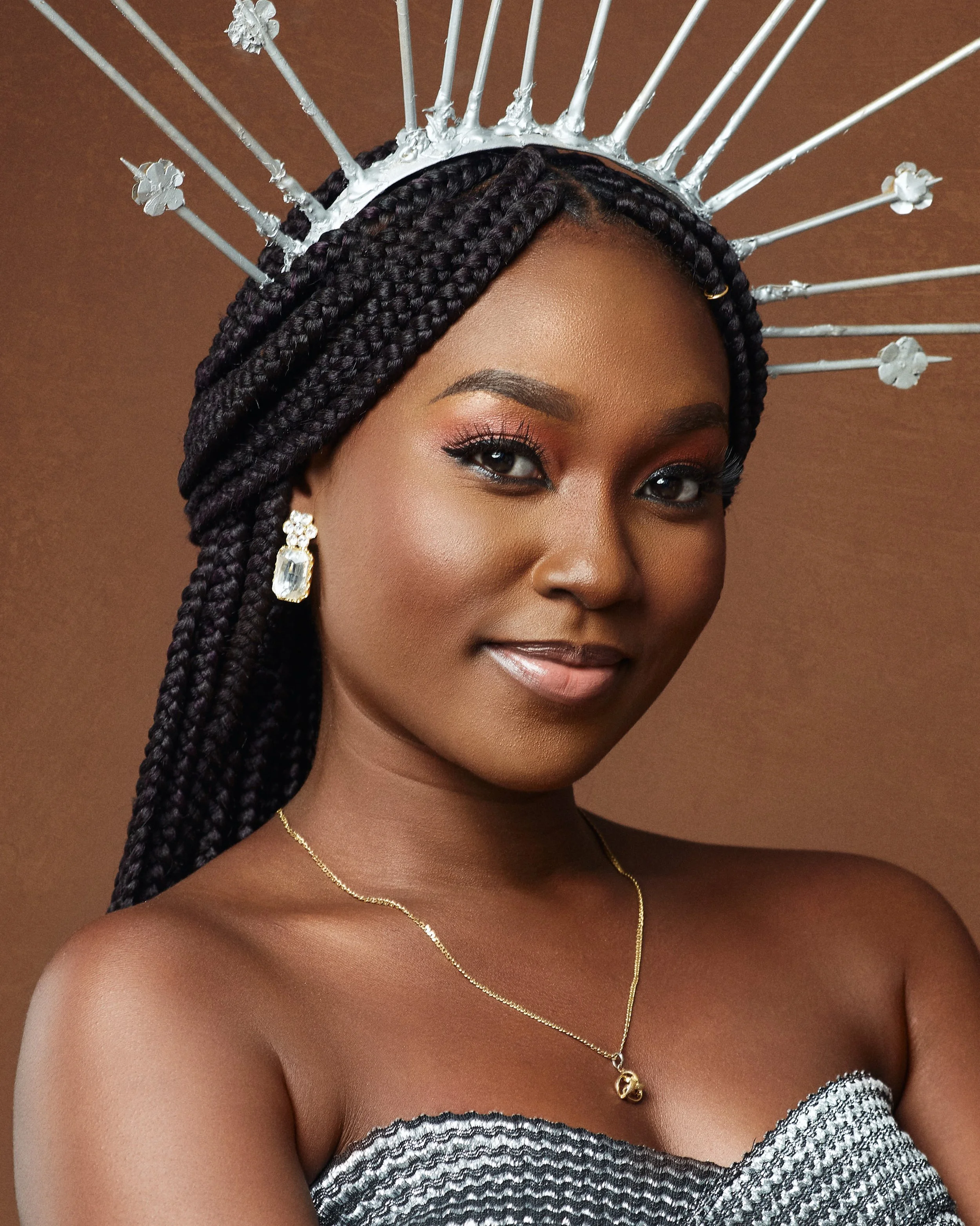 A close-up portrait of a woman with dark skin, short hairstyle, and elegant makeup. She is holding a yellow flower near her face and wearing large gold rings and earrings. The background is plain and light-colored.