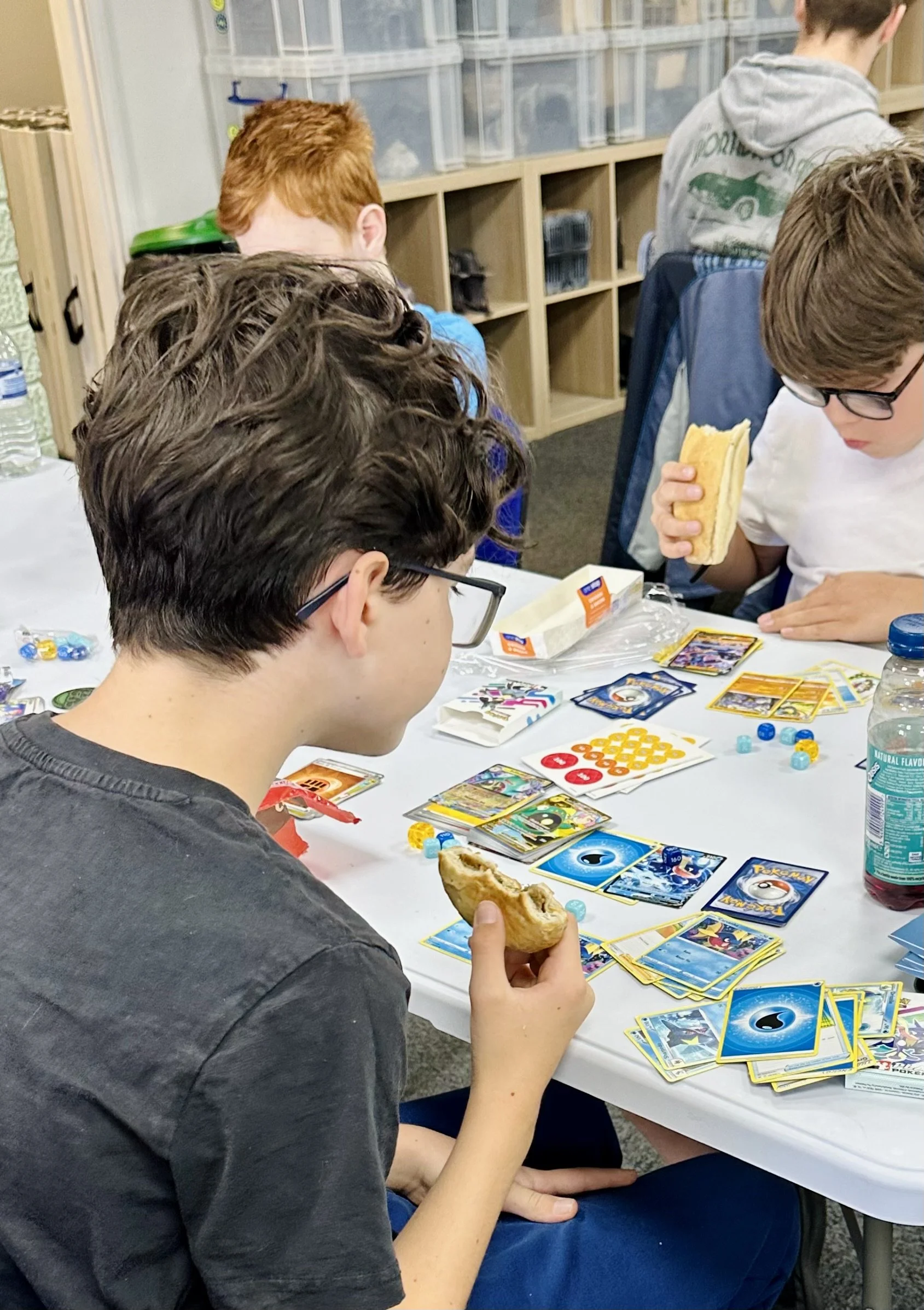 A group of kids playing a Pokémon trading card game at a table, with snacks and water bottles nearby.