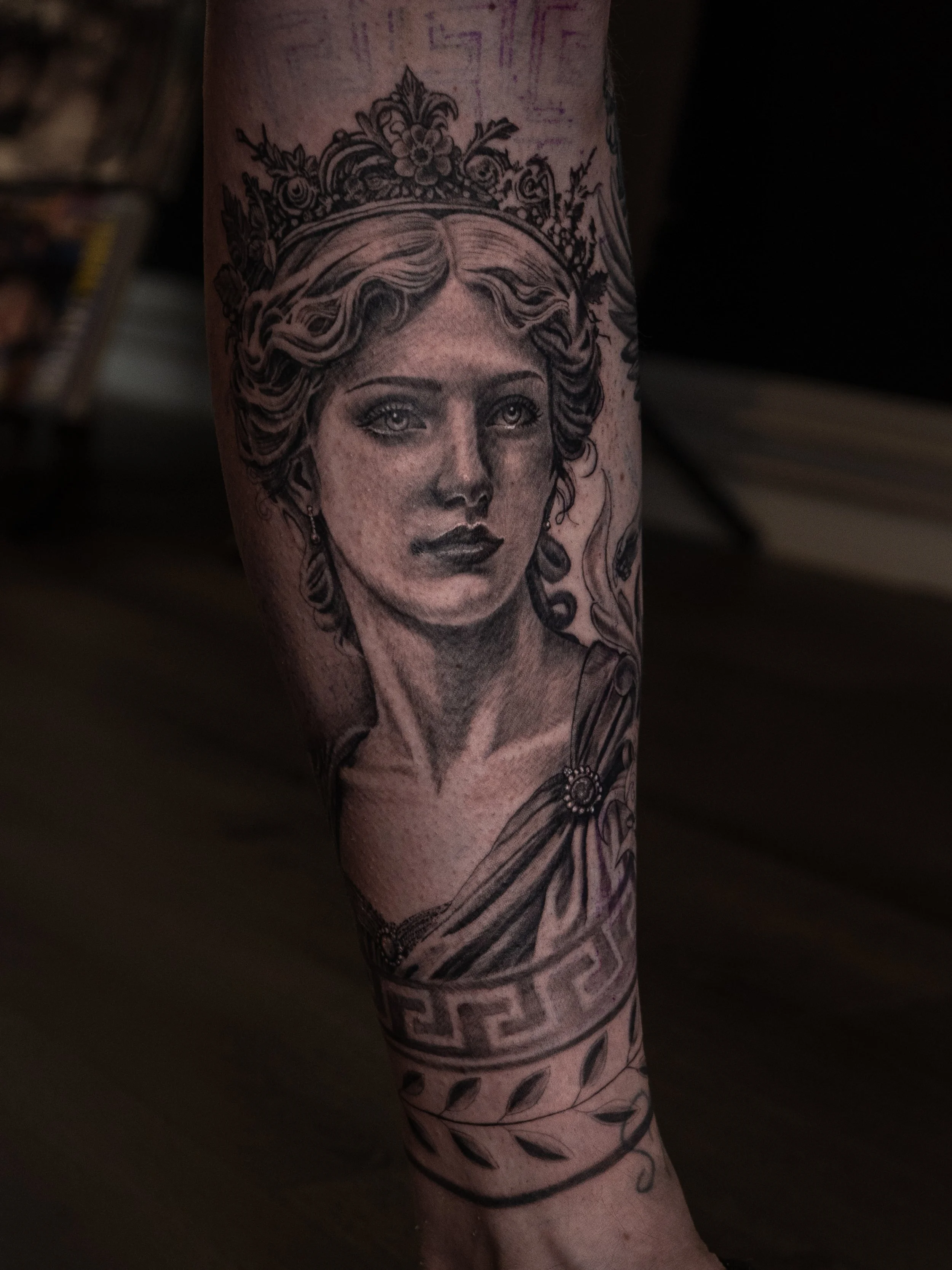 Tatuering av en kvinnlig gudinna med krona och draperi på armen.