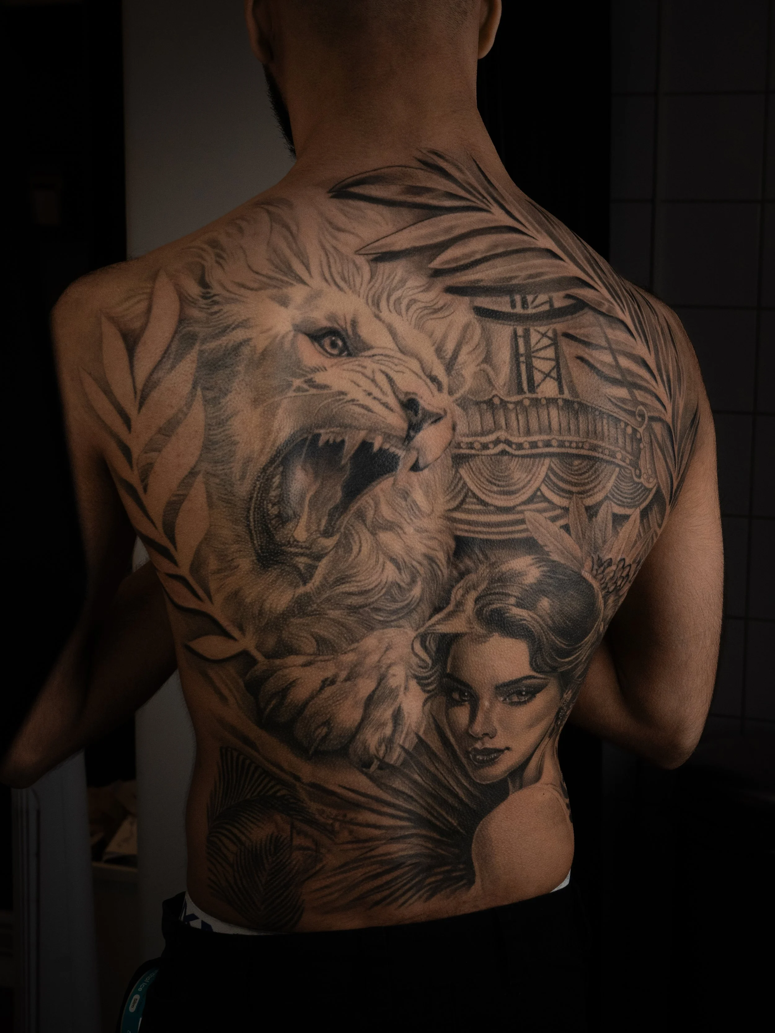 En person med ett stort svartvitt tatuering på ryggen som föreställer en lejon, en kvinna med mörkt hår, en stadssiluett och växtblad.