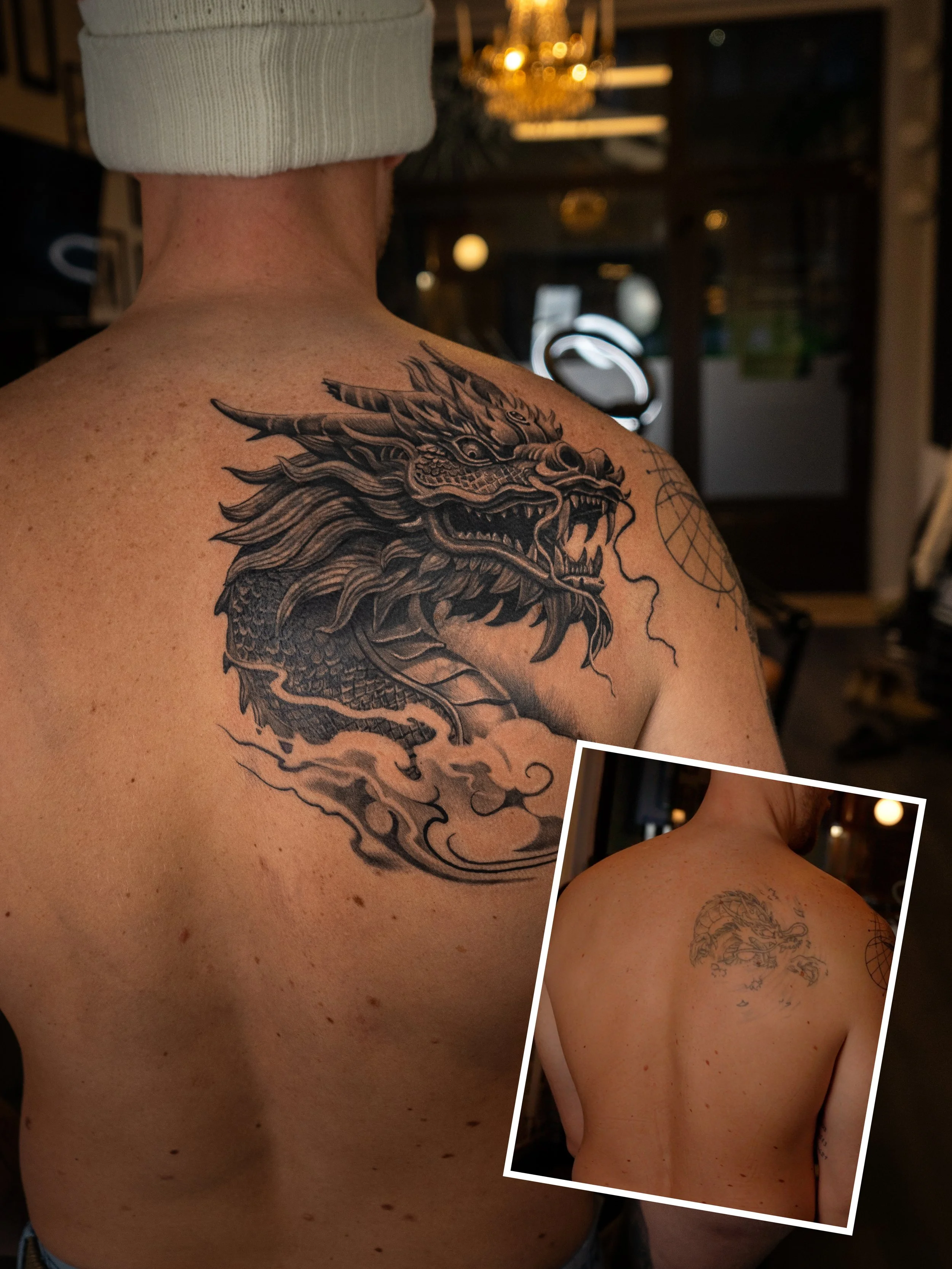 Person med en stor svart och grå drak-tatuering på vänster axel och övre ryggen, och en mindre skiss av samma drake på högra axeln, i ett inomhusmiljö.