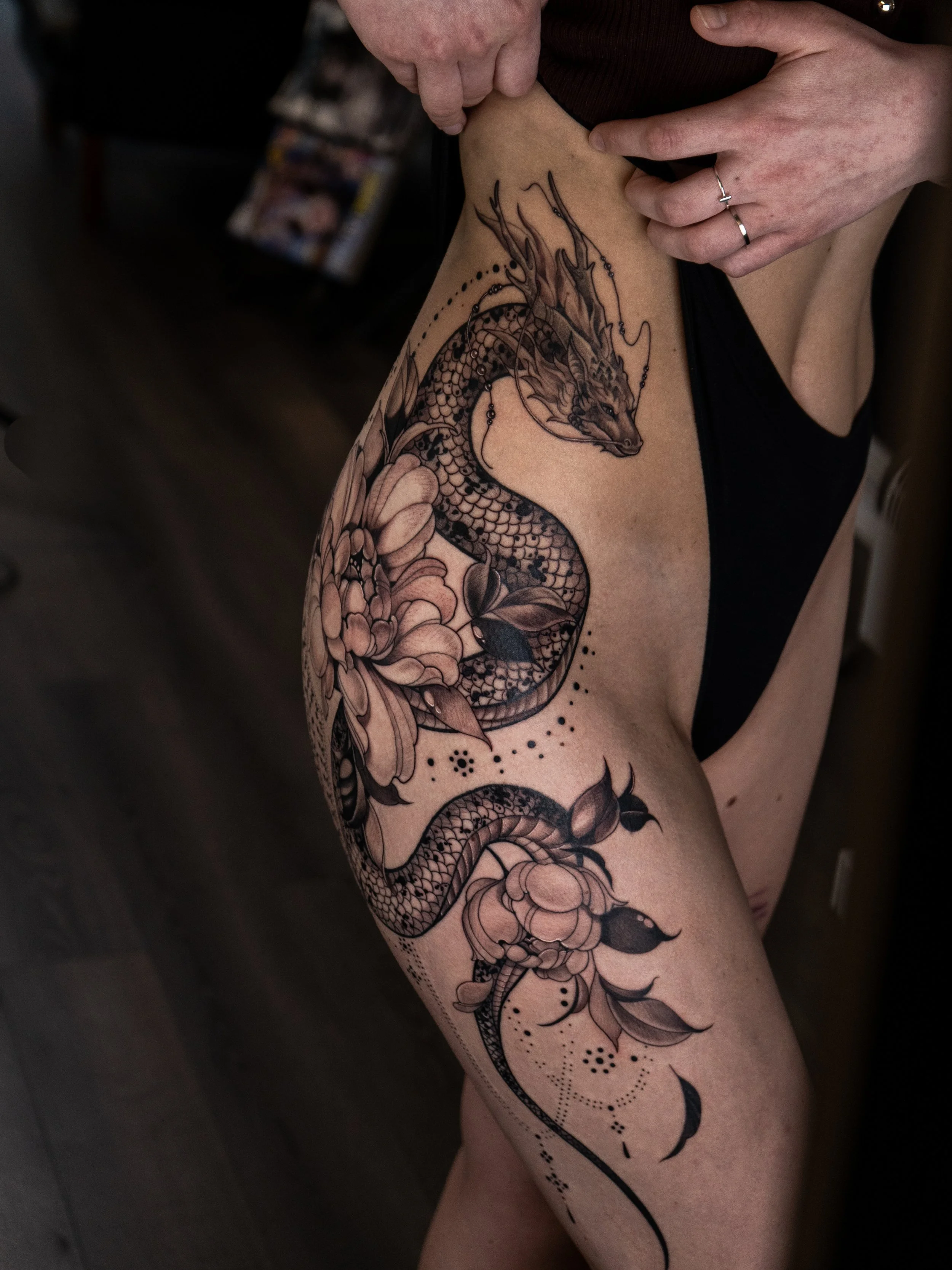 En person visar en stor, svart dekorativ tatuering på sin vänstra lår, som föreställer en drake och blommor.