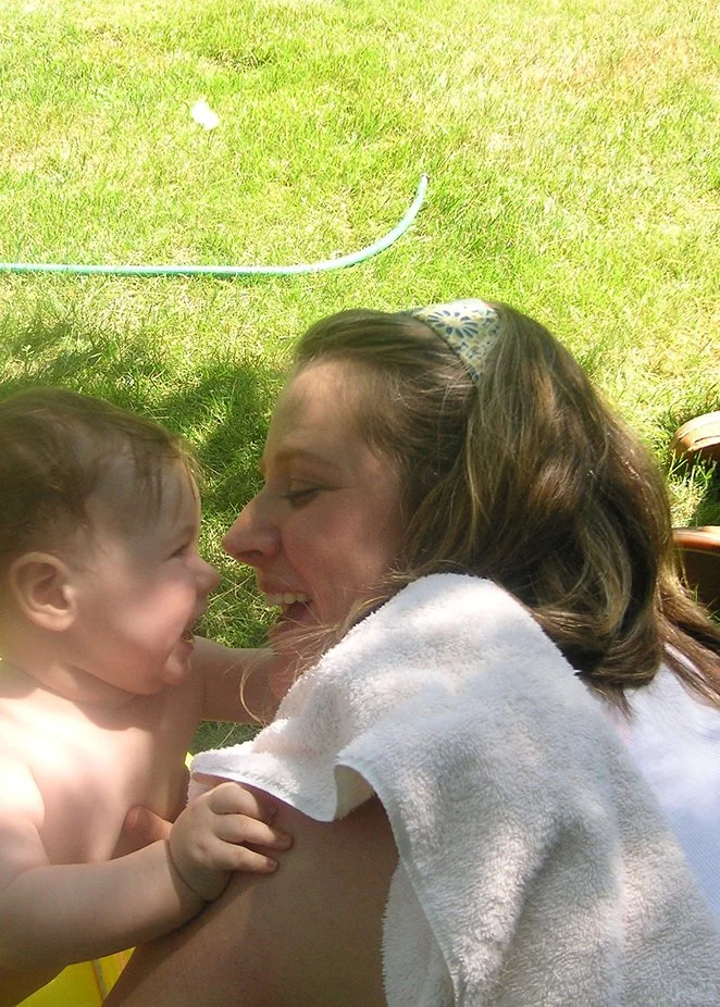 A woman and a young child smiling and leaning close together outside on a grassy lawn. The woman has a towel draped over her shoulder and is smiling at the child, who appears to be happy and playful.
