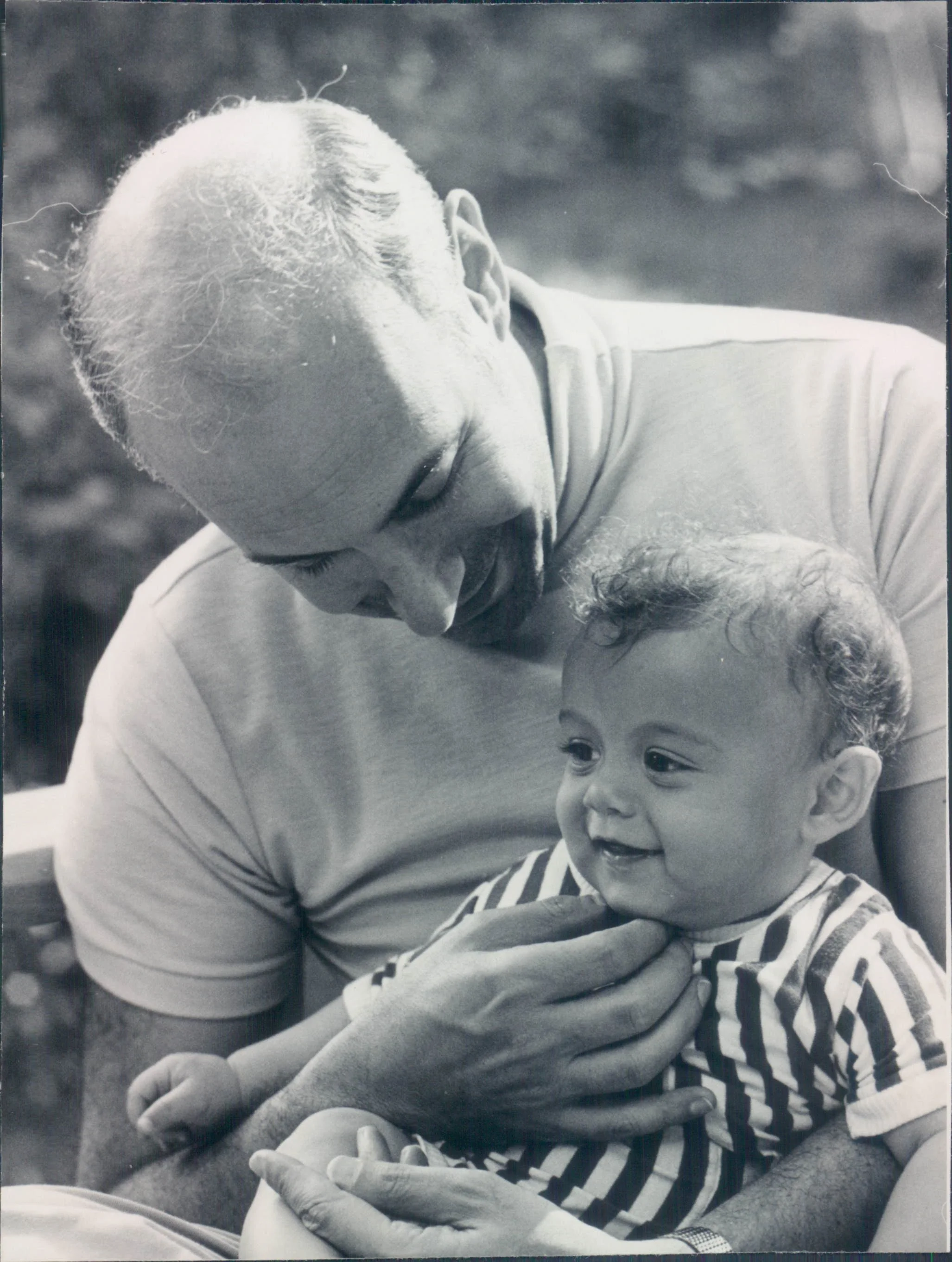 A black and white photo of an elderly man holding a young child, looking at each other affectionately, outdoors.