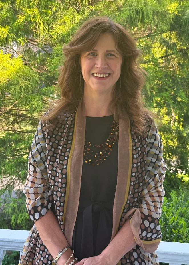 A woman with long brown hair smiling outdoors, standing in front of green trees, wearing a black top, polka dot cardigan, and layered necklace.