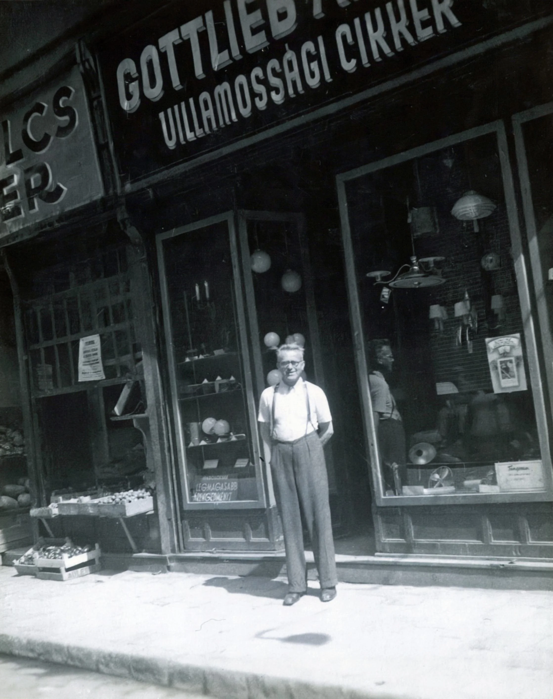 Black and white photo of a man standing outside a shopfront with large glass windows, displaying various items inside and signs in Swedish. The shop has a sign above that reads 'GOTTLIEDER VILLAMOSSAGICIRKEL'.