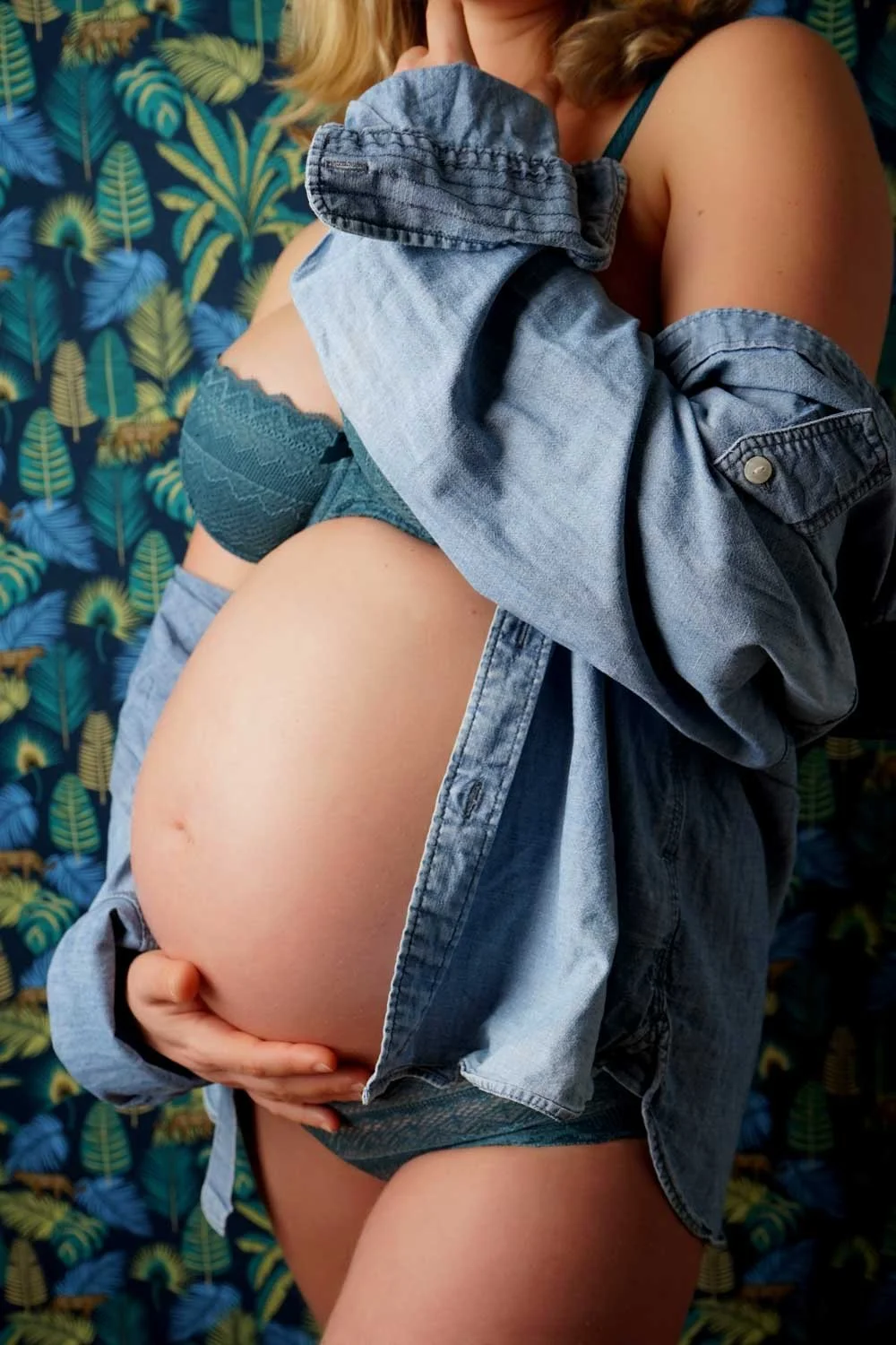 Ventre de fin de grossesse entre une chemise en jean ouverte et un décor de feuillages.