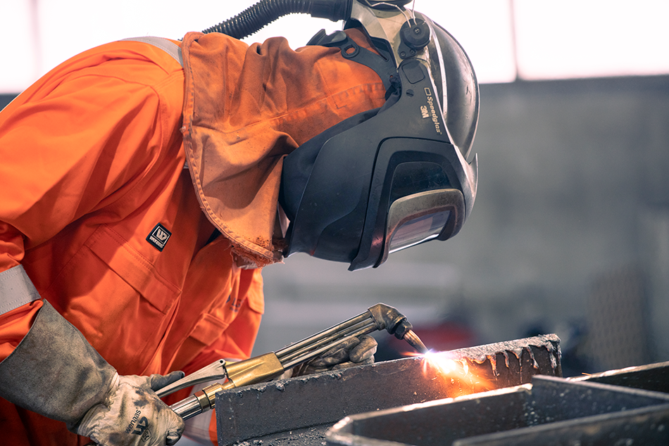 En person i orange arbeidsdress og vernebriller som sveiser metall.