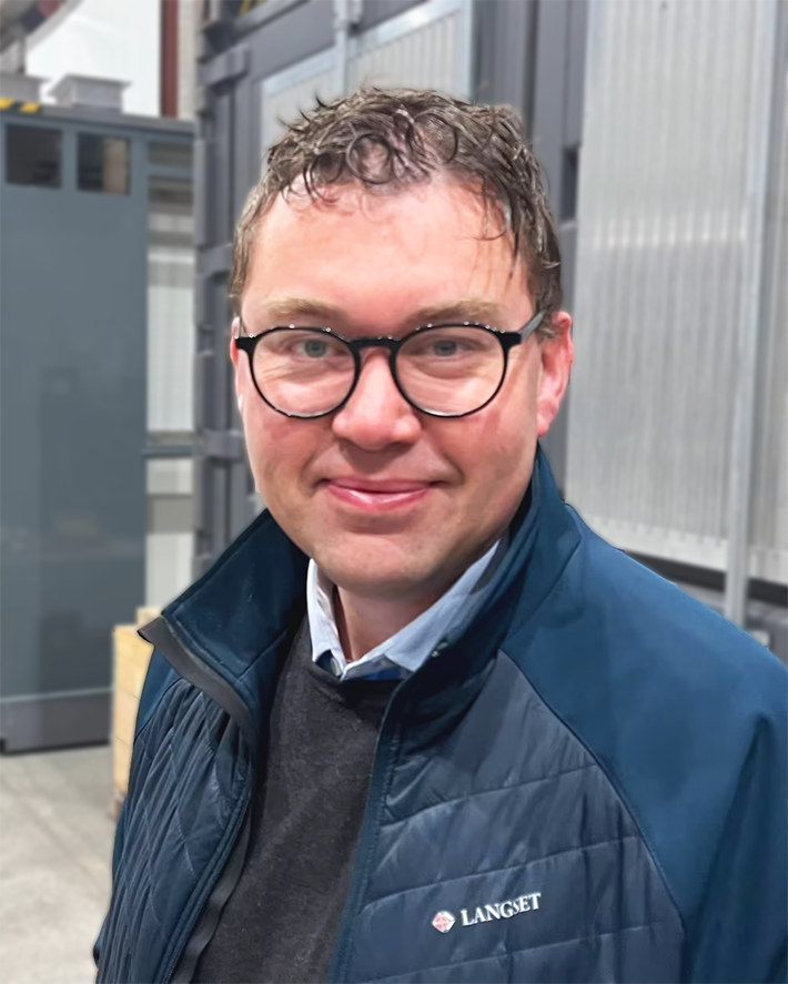 En mann med briller som smiler, iført en blå jakke i innendørs industriområde.