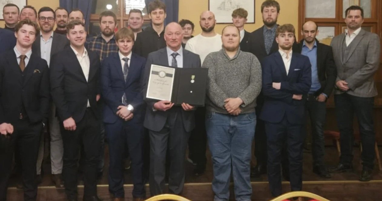 En gruppe menn i formelle klær, noen i dress, samles i et rom med gule vegger og bilder på vegg, en av dem holder en diplom i hånden, en pris Langset fikk for inkludering og mangfold.