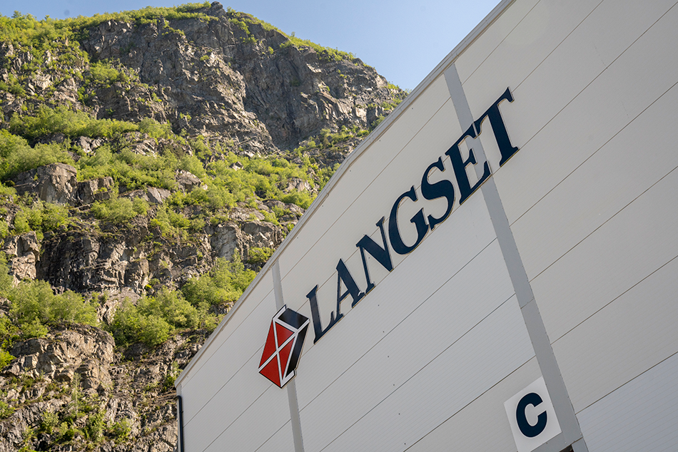 Nærbilde av en fabrikkbygning med Langset-logo, omgitt av fjell og grønne trær under klar himmel.