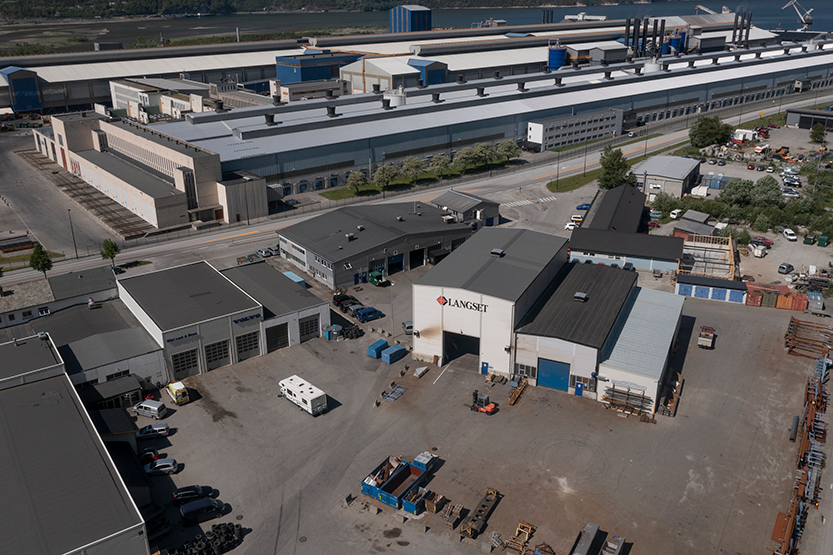 Utsikt over en industriell bedrift med lagerbygninger, containere og utstyr, med vann i bakgrunnen.