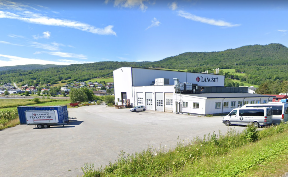 Utvendig bilde fra Langset Mek i Eidsvåg. En hvit verkstedbygning og en stor hall, med grønn plen i forgrunnen og fjell, blå himmel og sol i bakgrunnen.
